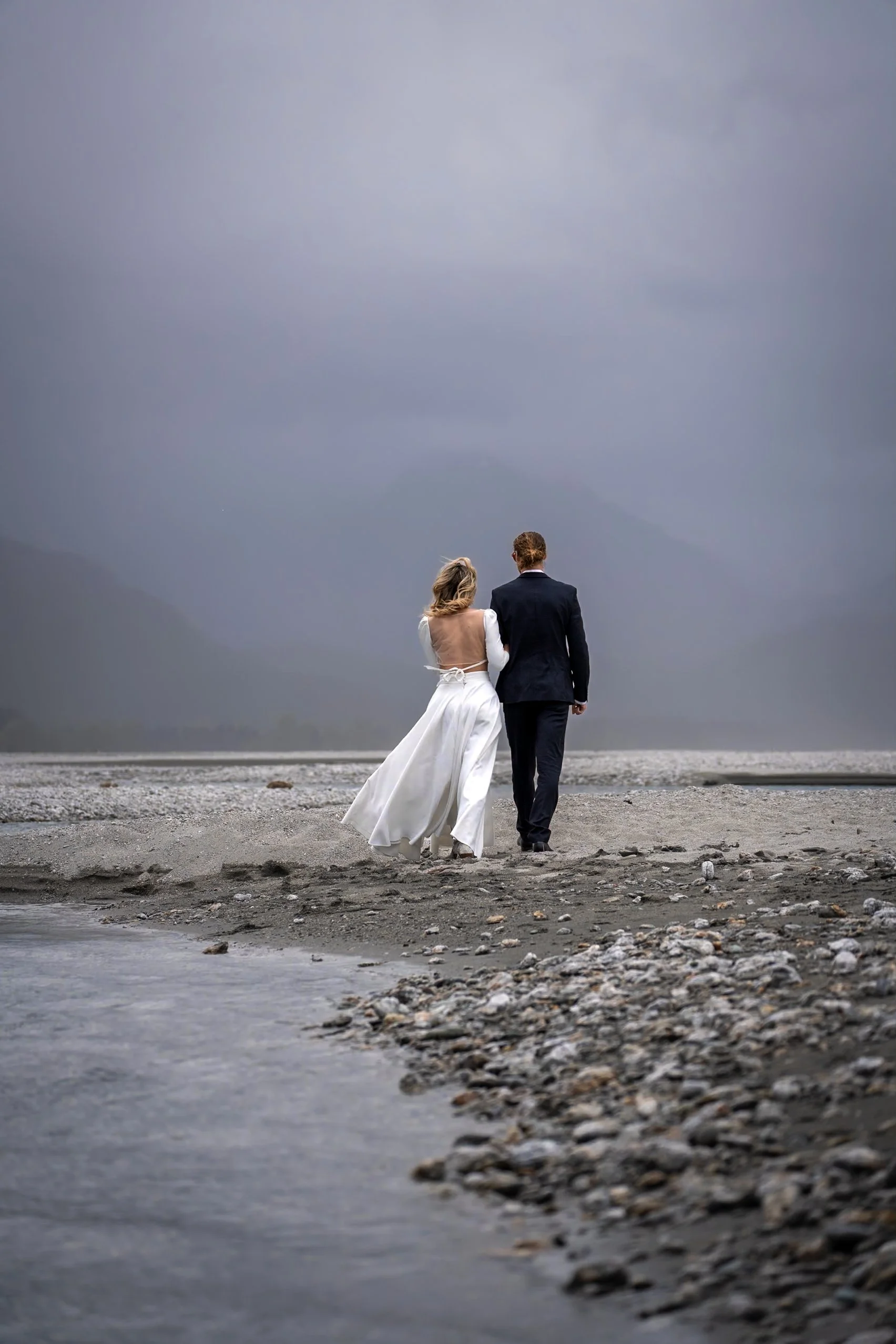 A bride and groom walking together on a rocky beach under a cloudy sky, with the bride in a white wedding dress and the groom in a black suit.
