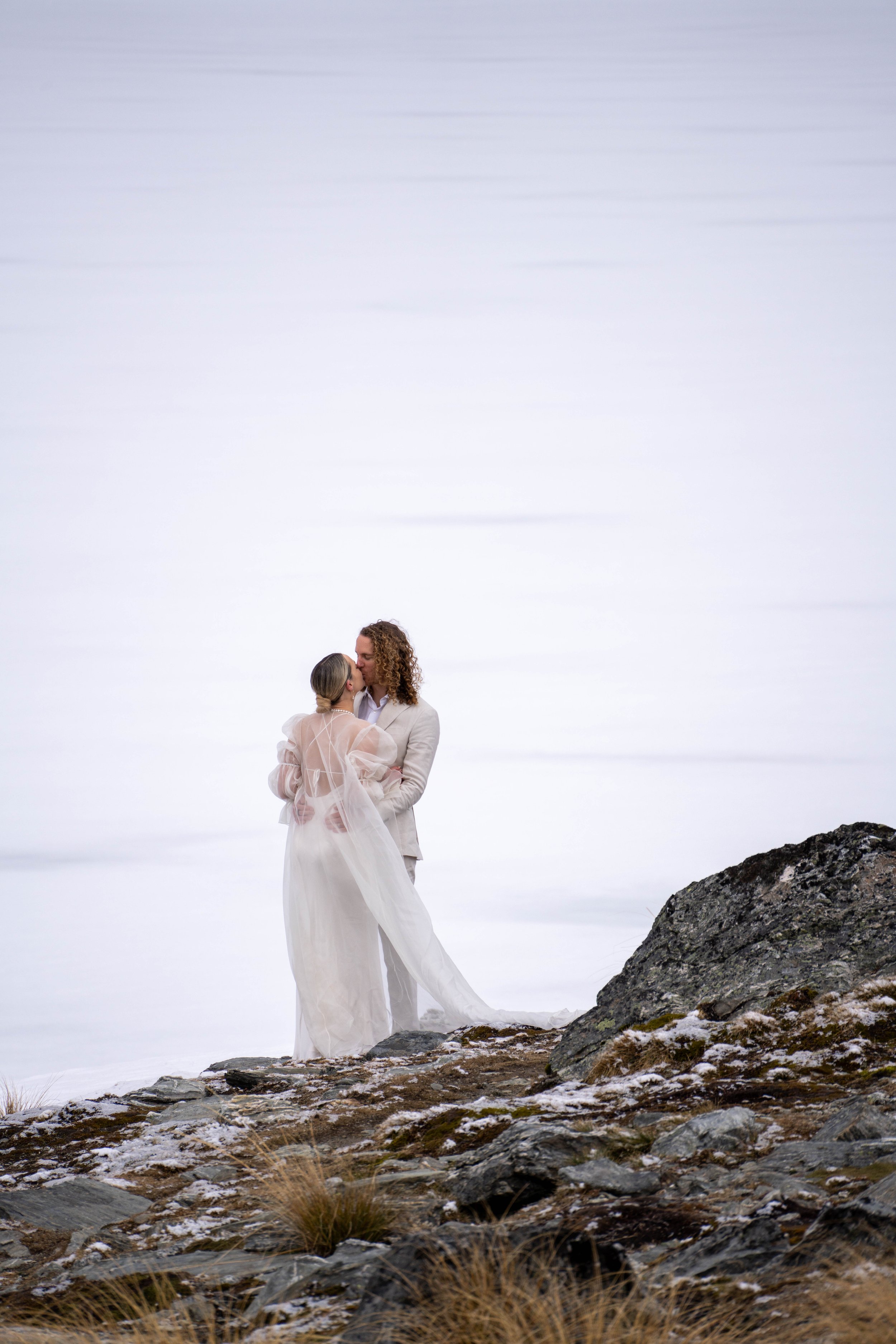A couple dressed in wedding attire sharing a kiss on a rocky, snow-covered landscape with a mostly white and cloudy sky in the background.