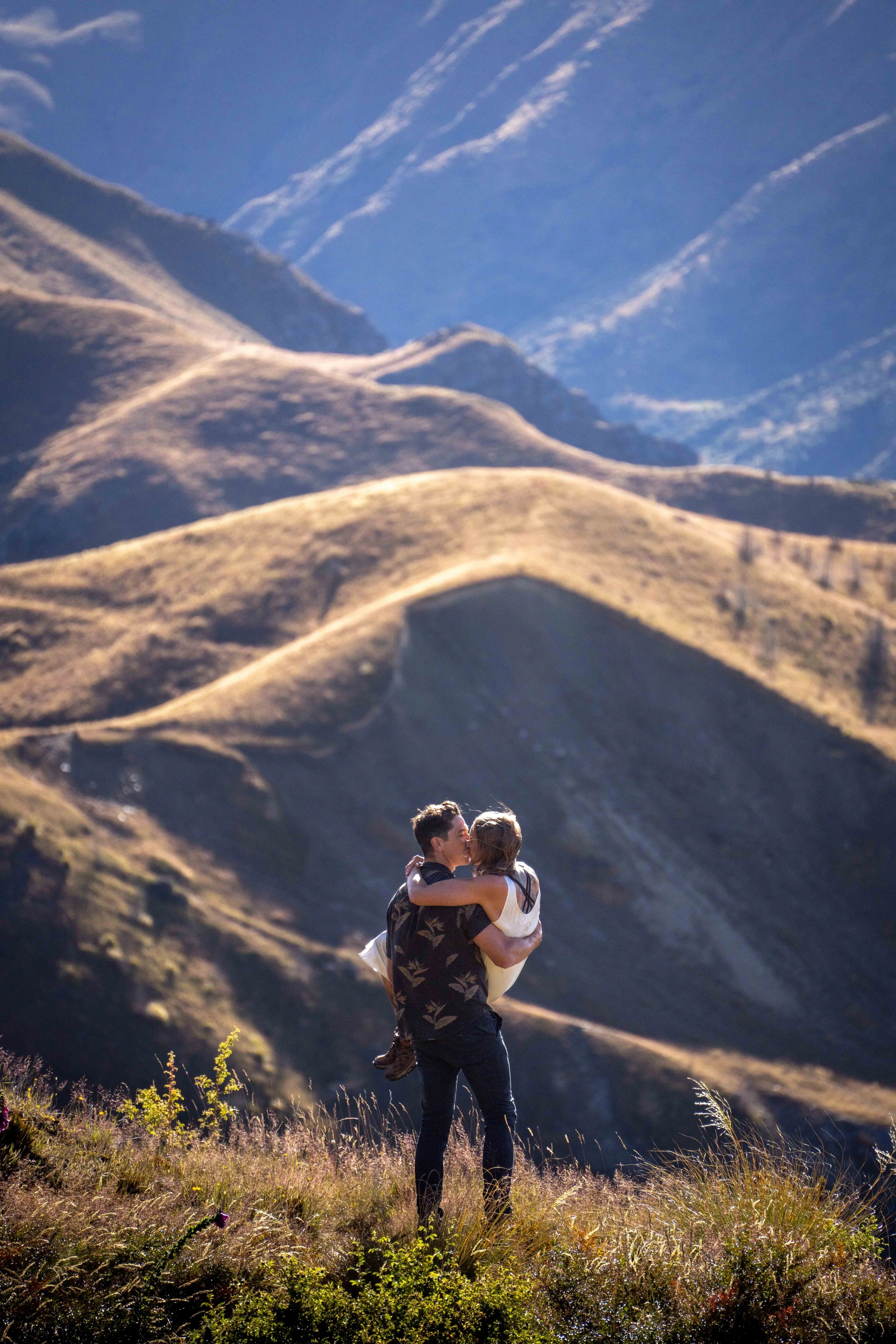 A man carrying a woman in his arms in a mountainous landscape with rolling hills and a blue sky.