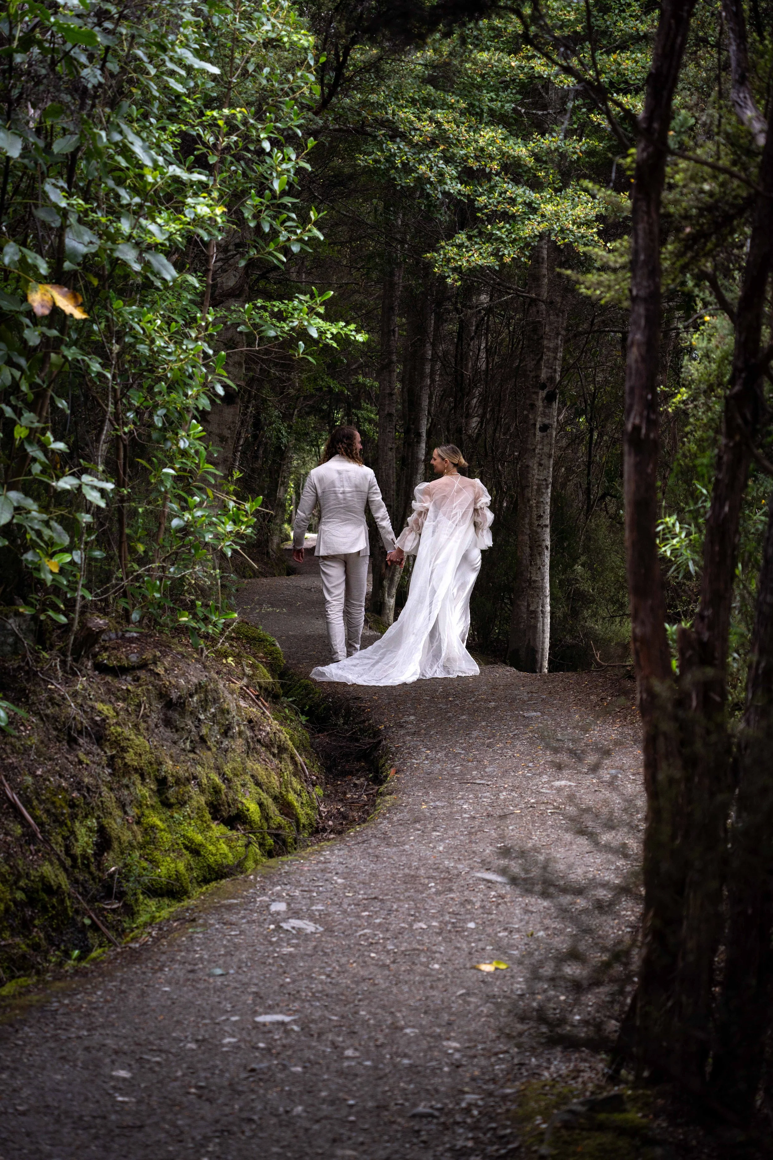A bride and groom holding hands walk along a forest trail. The bride wears a white wedding dress with long train, and the groom wears a light-colored suit.