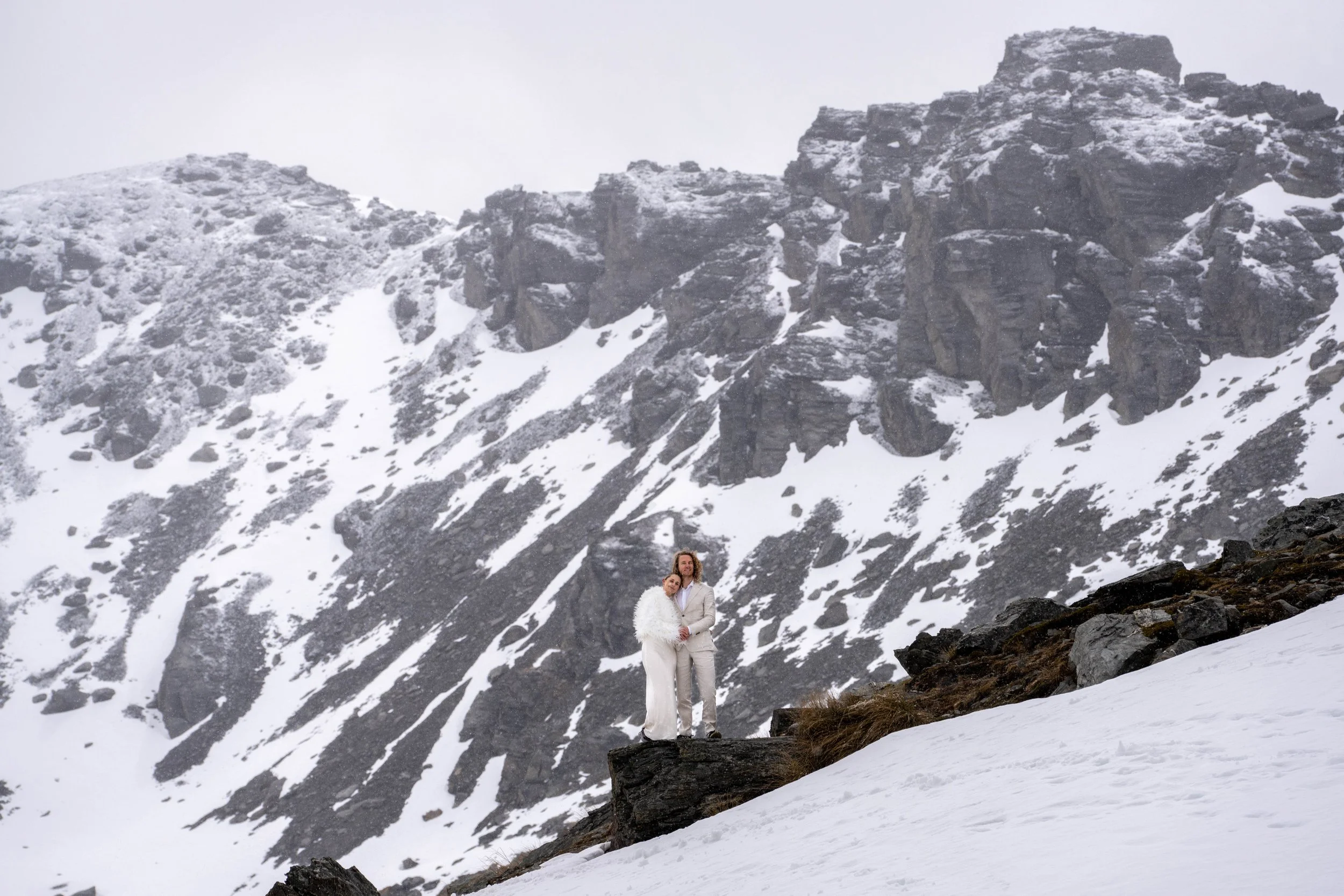 Couple dressed in white standing close together on a snowy mountain slope with a rocky mountain range in the background.