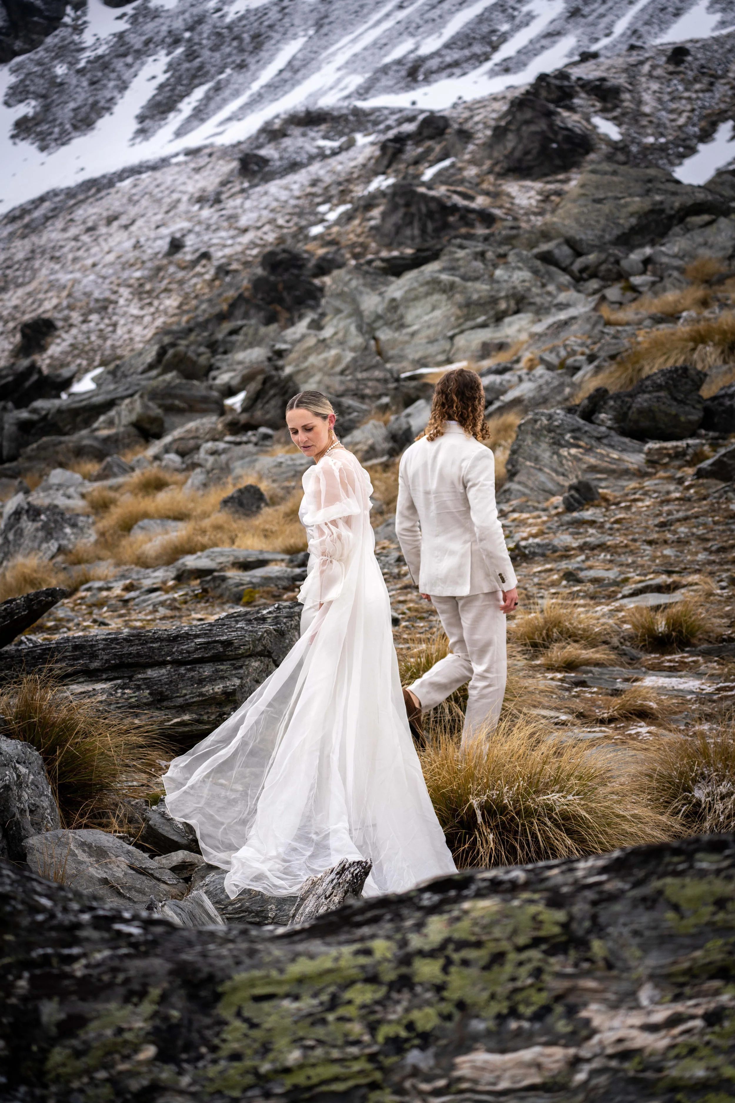 A bride in a white wedding dress and a groom in a white suit stand in a rocky, mountainous landscape with snow patches.