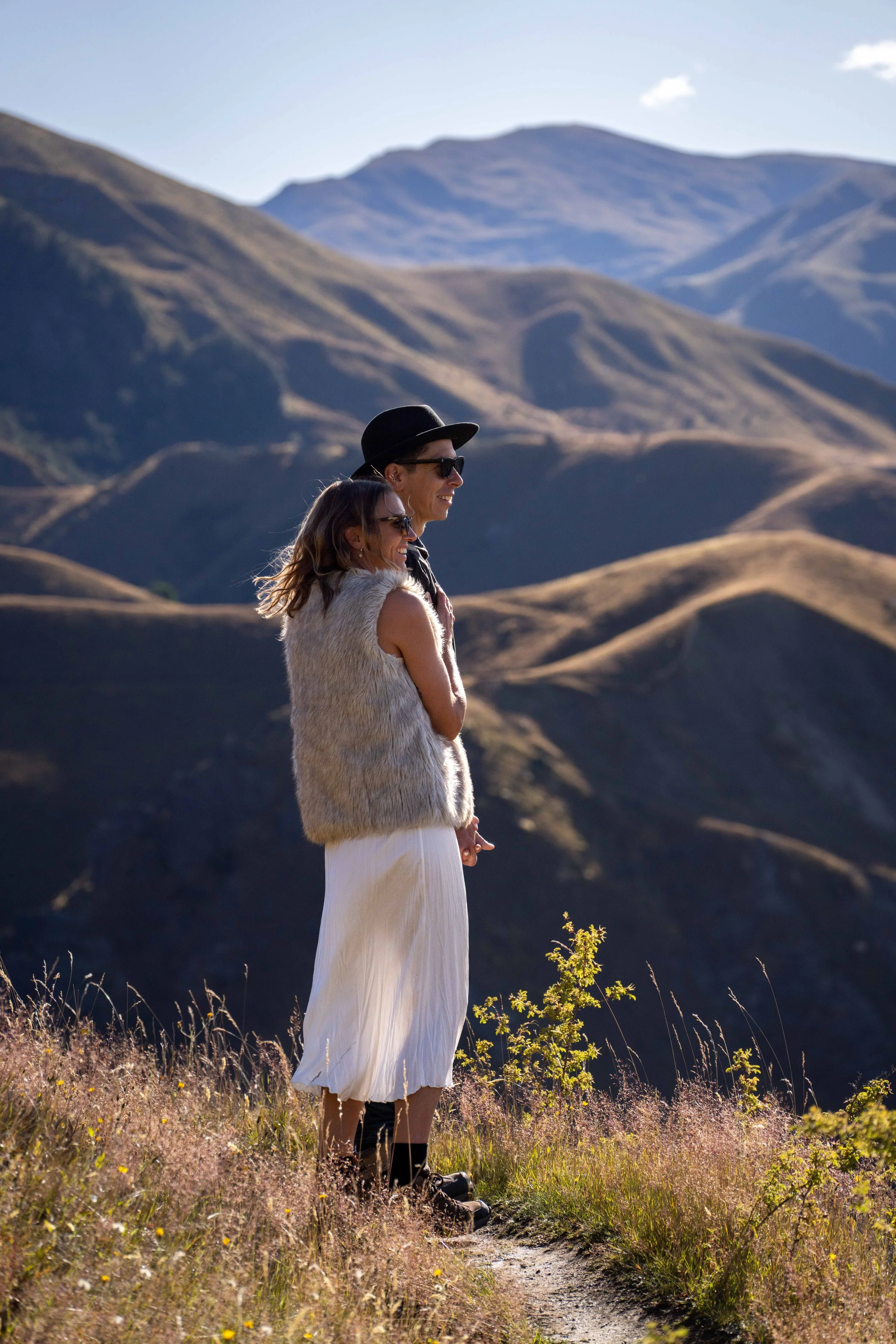 A man and woman standing on a trail in a mountainous landscape, smiling and enjoying the view in bright sunlight.