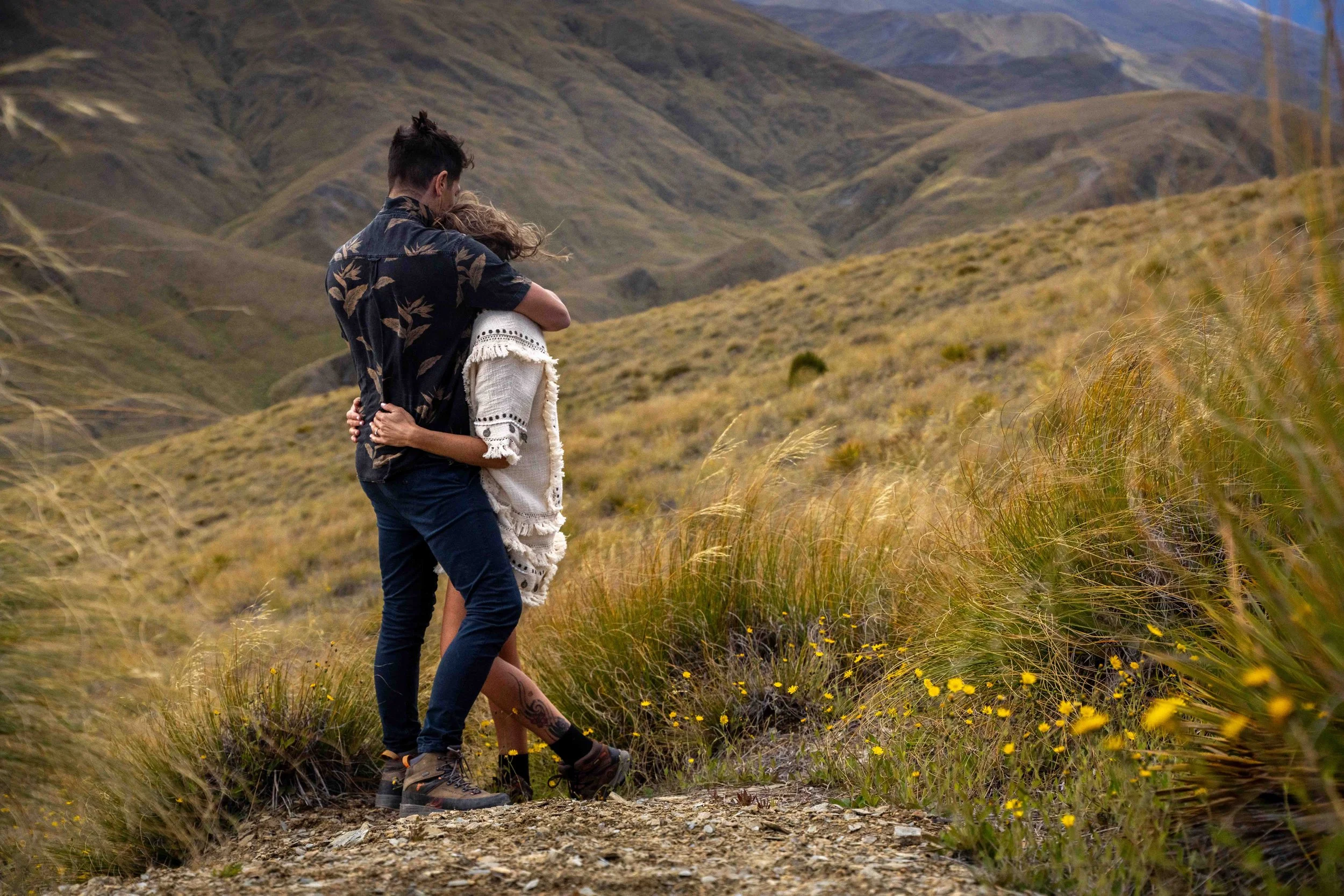 A couple embracing on a trail in a mountainous landscape with grassy hills and yellow wildflowers.