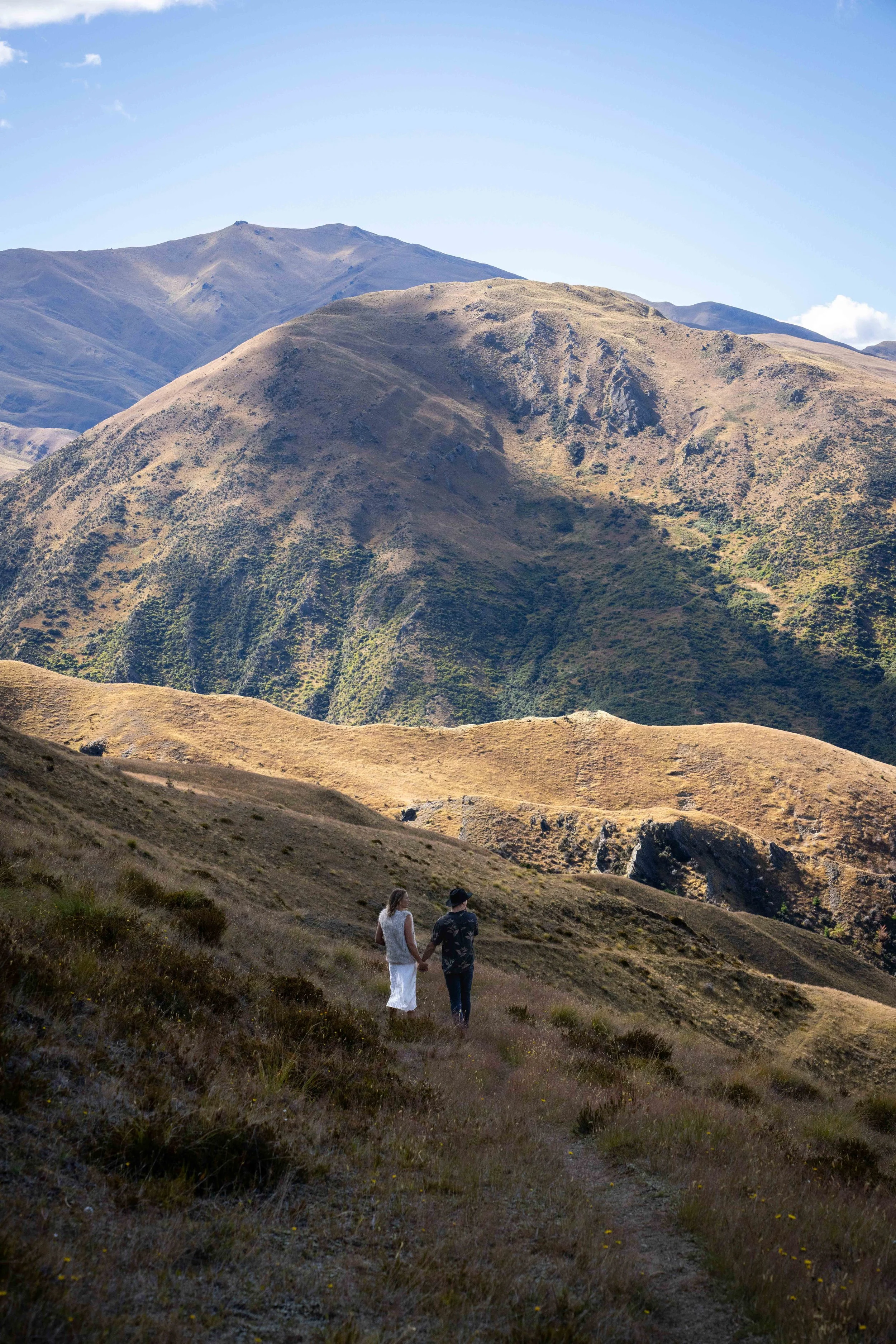 Two people holding hands walking on a mountain trail with large, dry mountains in the background under a blue sky.