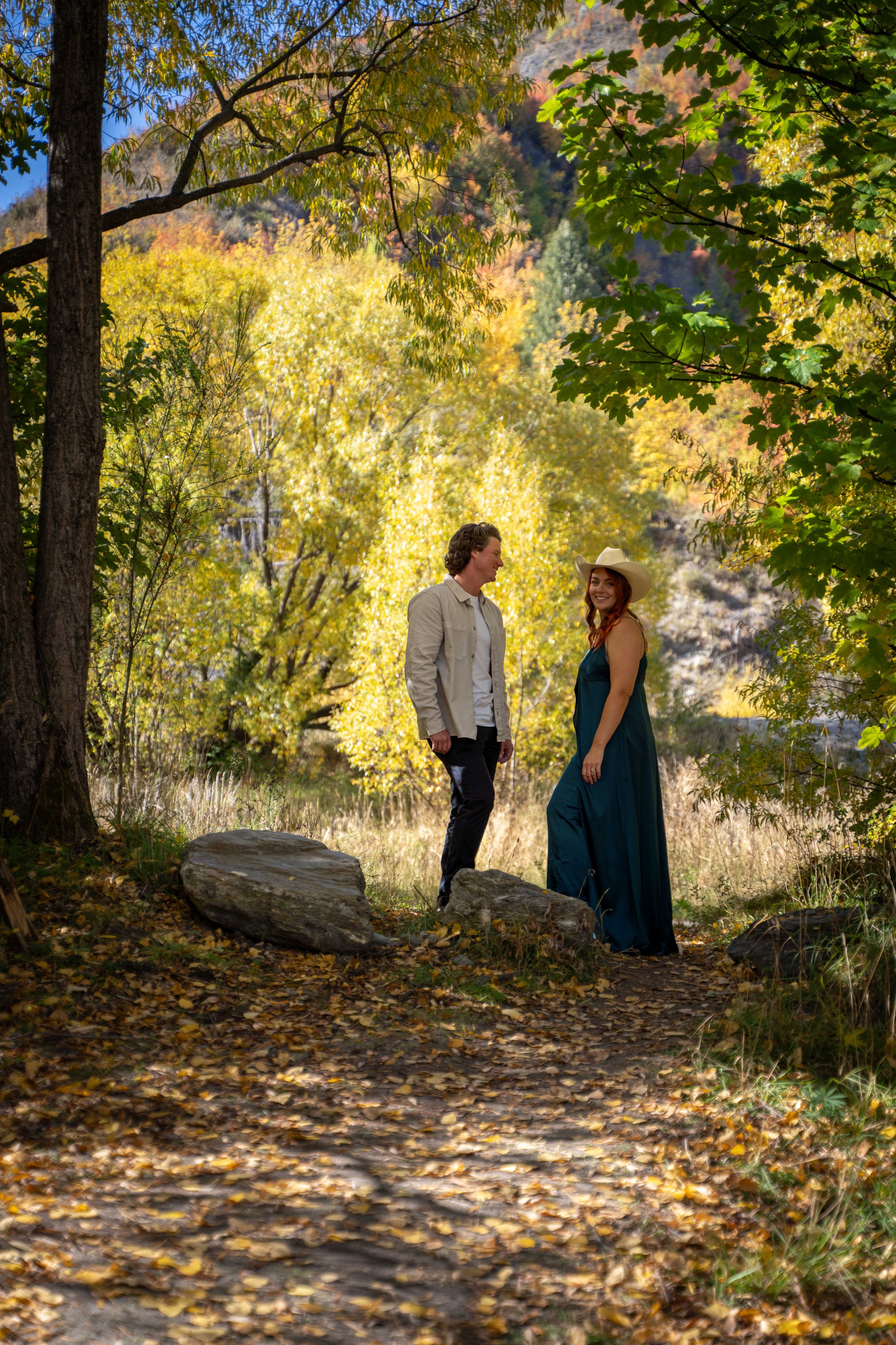 A couple standing on a forest trail surrounded by colorful autumn trees, with rocks on the path and sunlight filtering through the leaves.