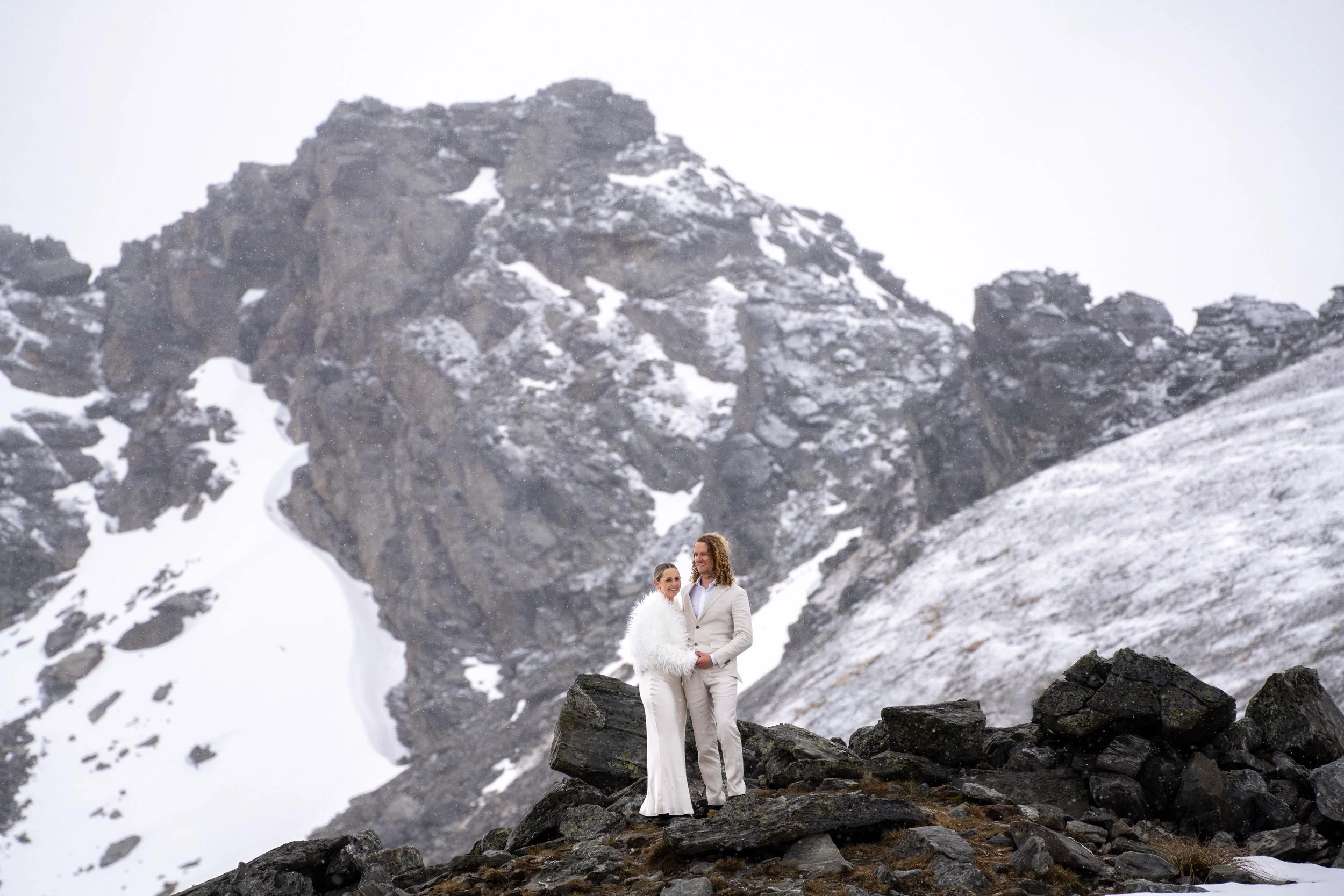 A couple dressed in white standing on rocky terrain with snow-covered mountains in the background.