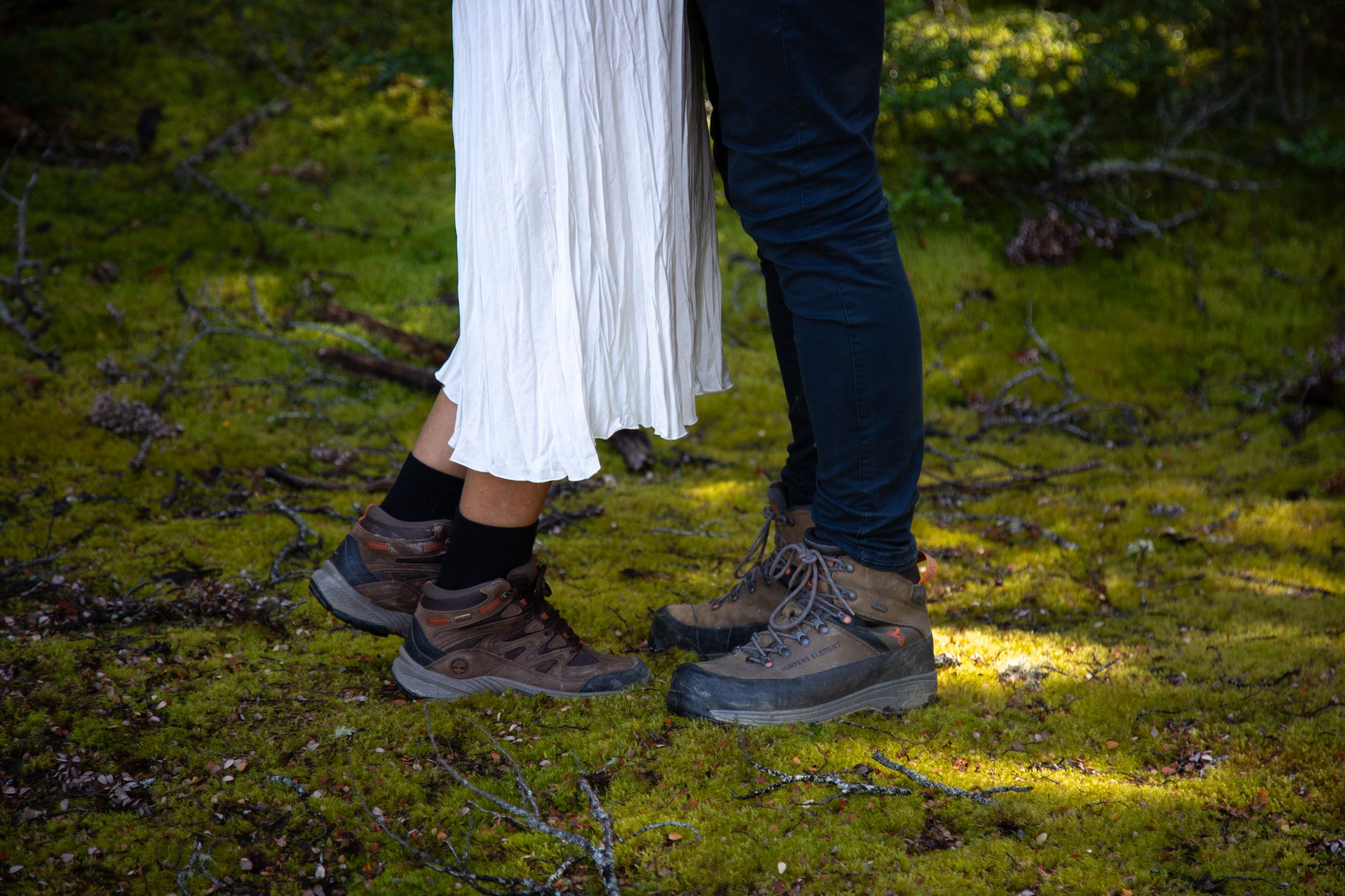 Close-up of two people standing on green mossy ground in a forest, wearing hiking boots and wedding attire, partially visible from the knees down.