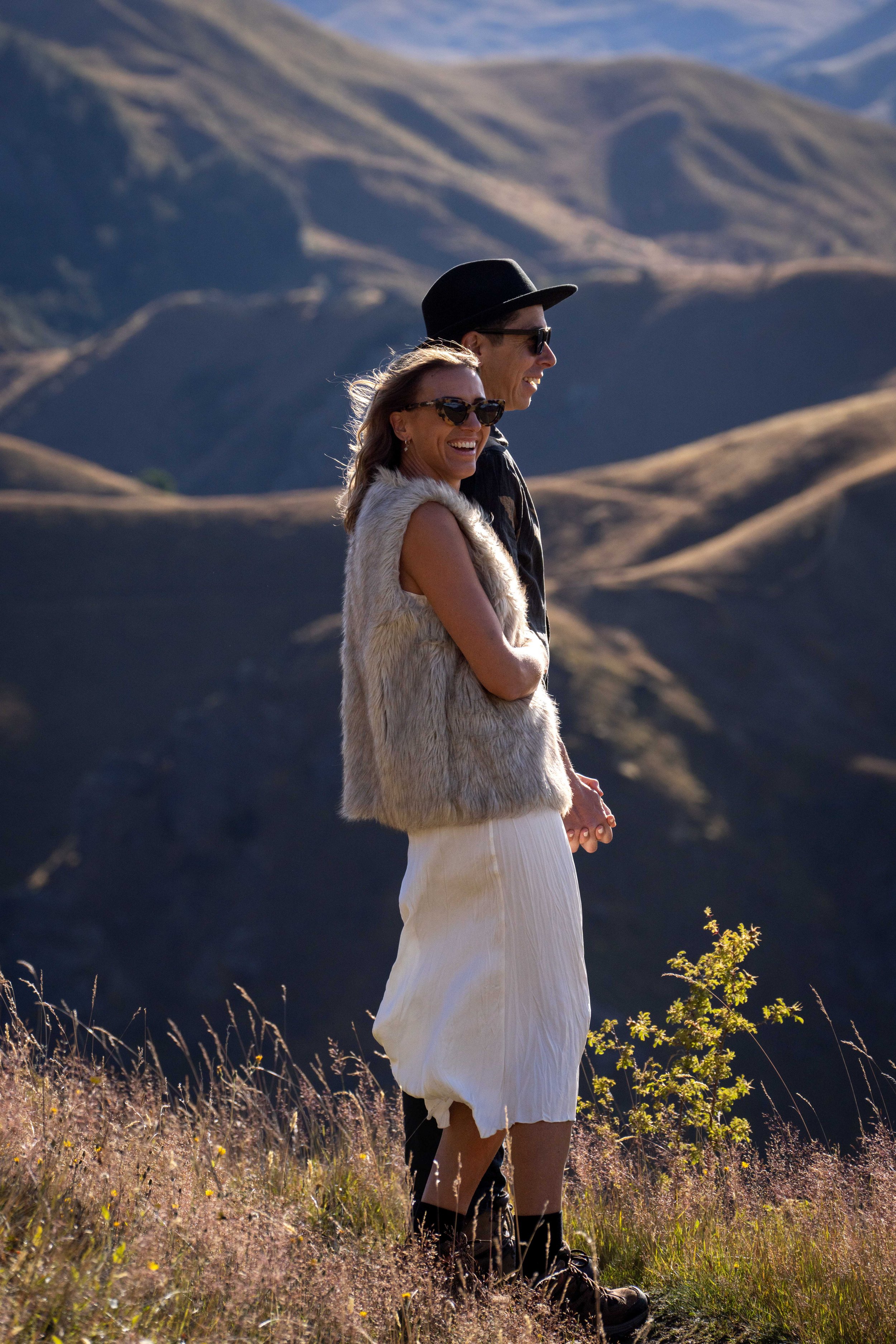 A smiling woman and man standing in a field with mountains in the background, enjoying a sunny day.