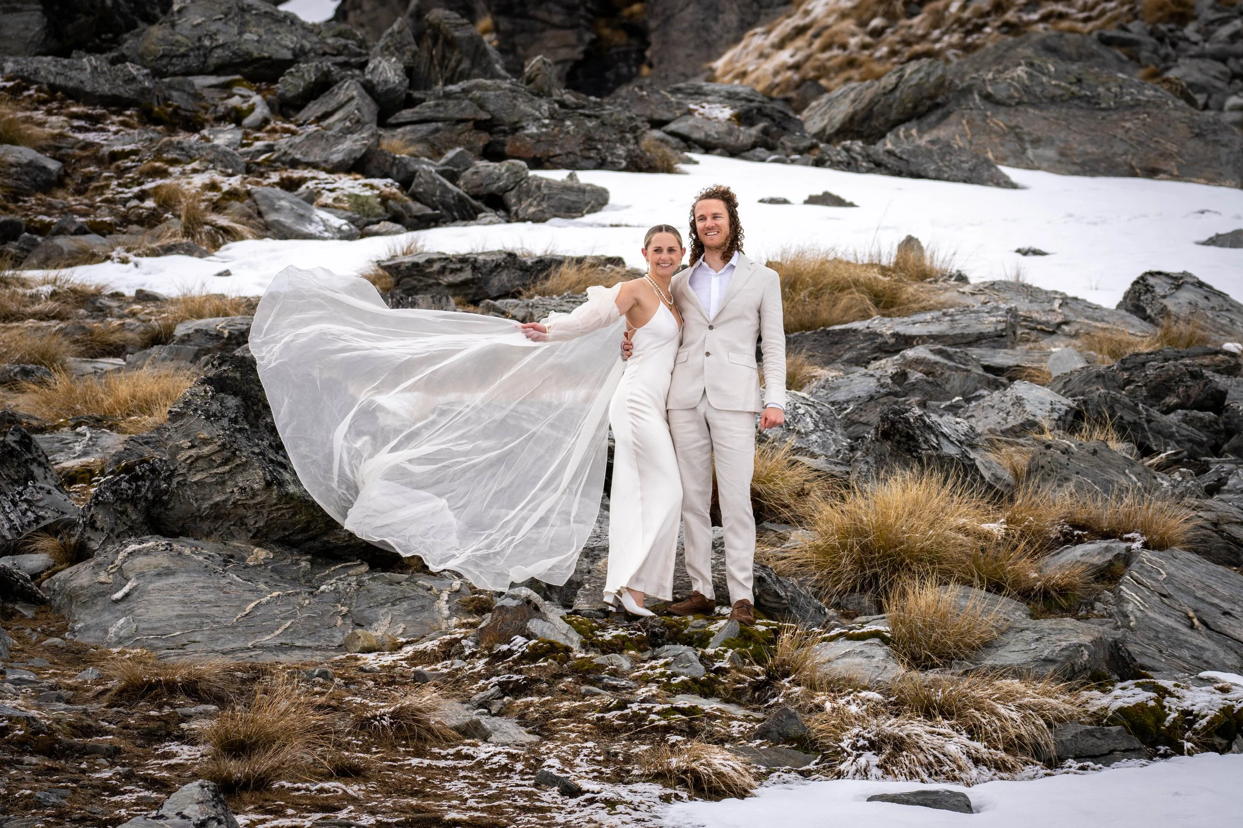 A couple in wedding attire standing on rocky terrain with patches of snow and dry grass, smiling at each other.