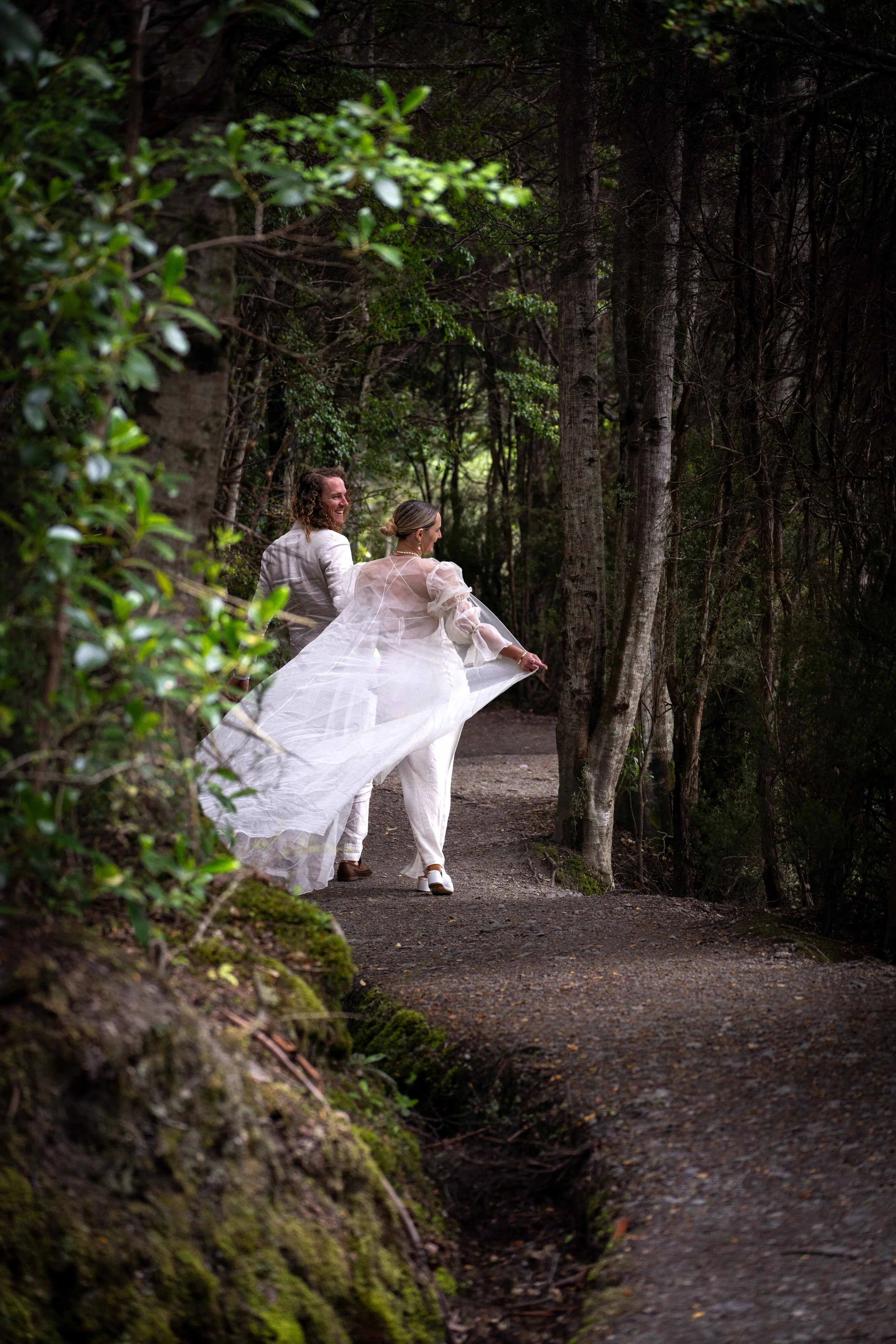 Two brides walking on a forest pathway, one in a white dress and the other in a light-colored suit, smiling and holding the train of the bride's wedding dress.