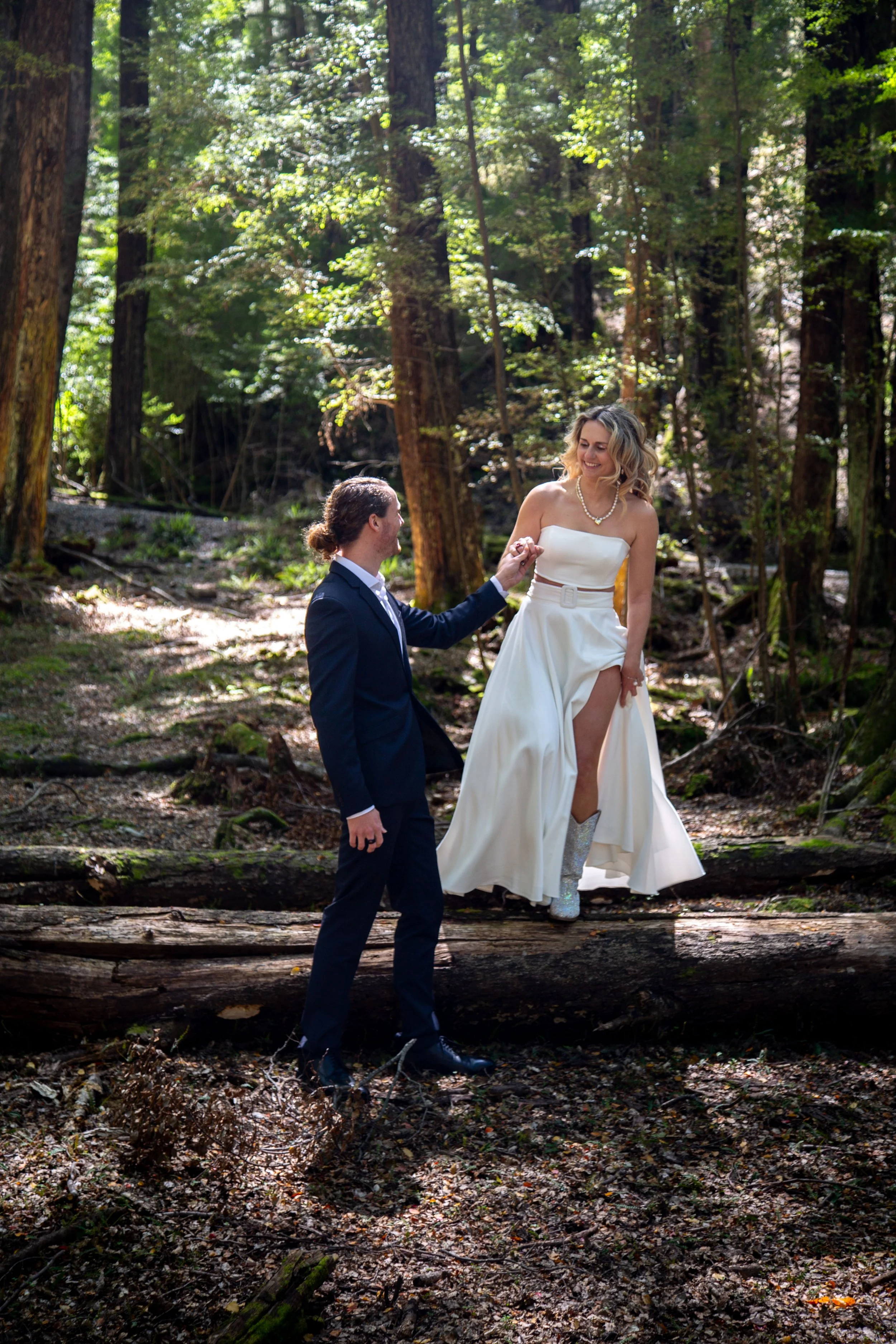 A bride and groom in wedding attire in a forest, with the bride standing on a log and the groom holding her hand.