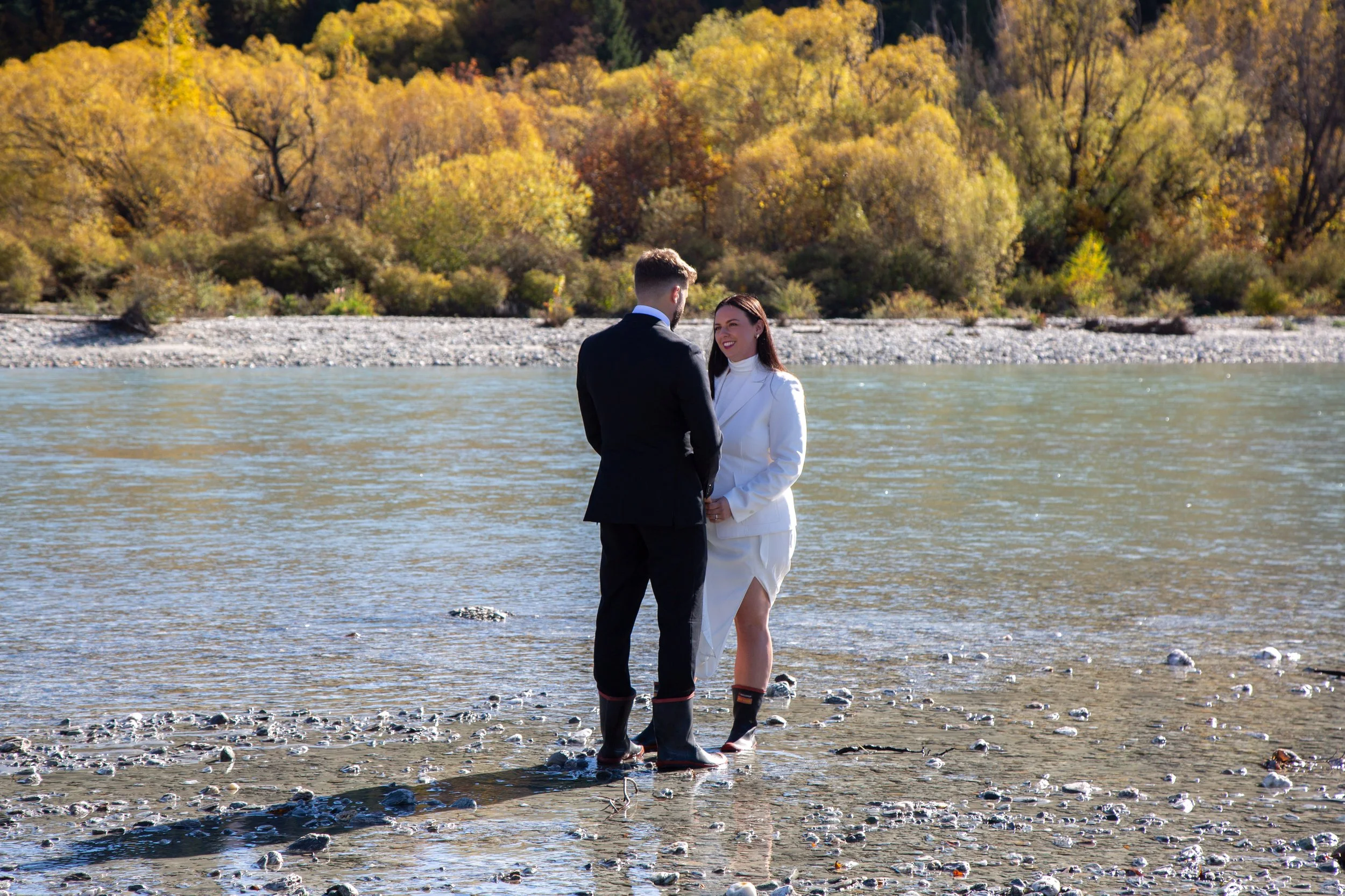 A couple dressed in formal attire standing in shallow water near a riverbank, surrounded by autumn trees with yellow and orange leaves.
