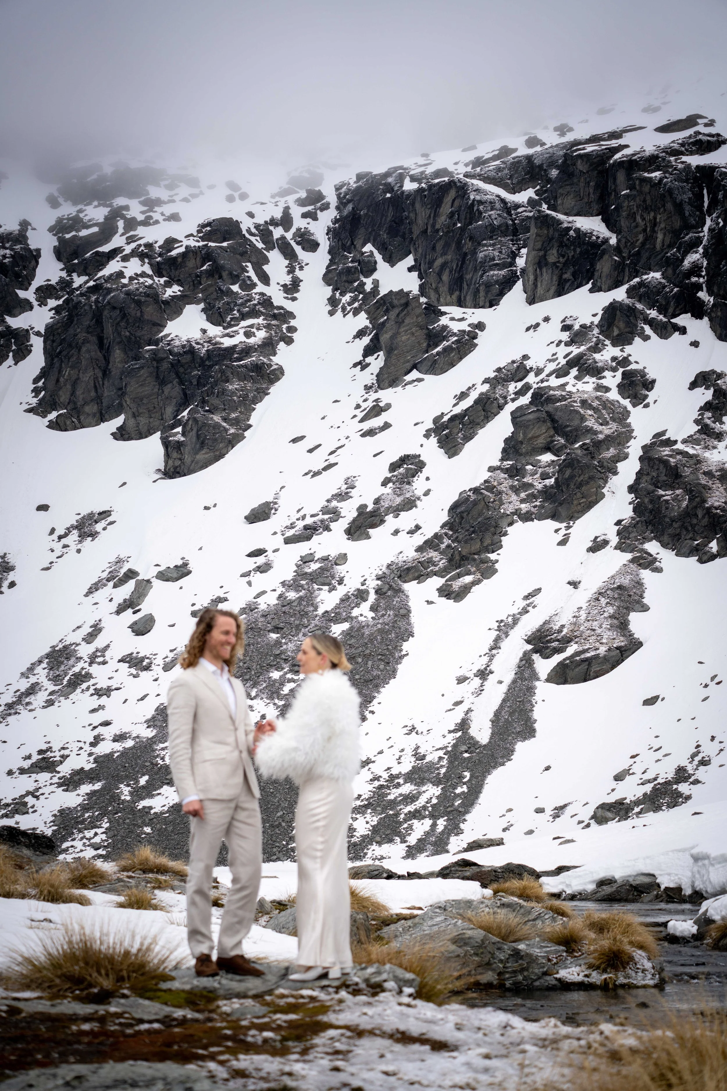 A couple dressed in formal white attire standing in a snowy mountain landscape with large rocks and a flowing stream.