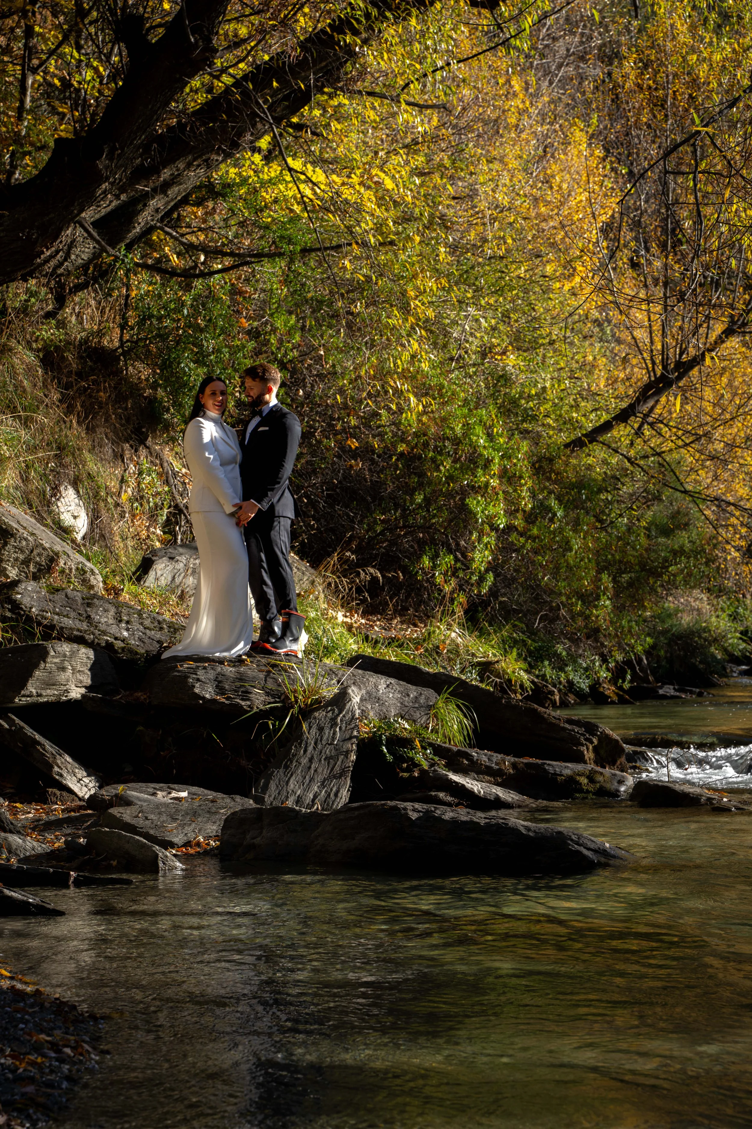 A couple in wedding attire standing on rocks by a creek in a forested area with colorful autumn leaves.