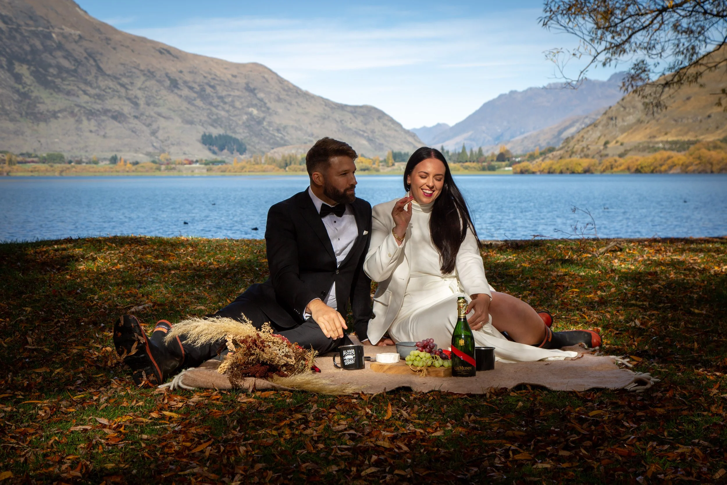 A couple dressed in formal and casual attire having a picnic by a lake with mountains in the background. The woman is smiling while holding a snack, and the man is seated beside her, pointing towards the woman.