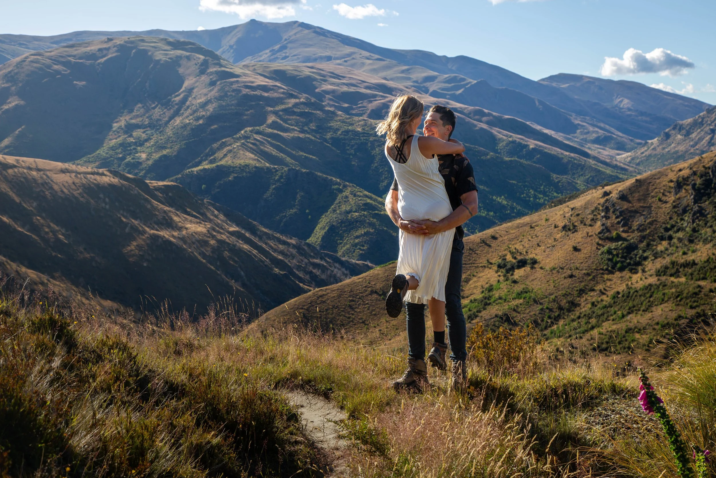 A man lifting and embracing a woman in a white dress in a mountainous landscape with green hills and clear blue sky.