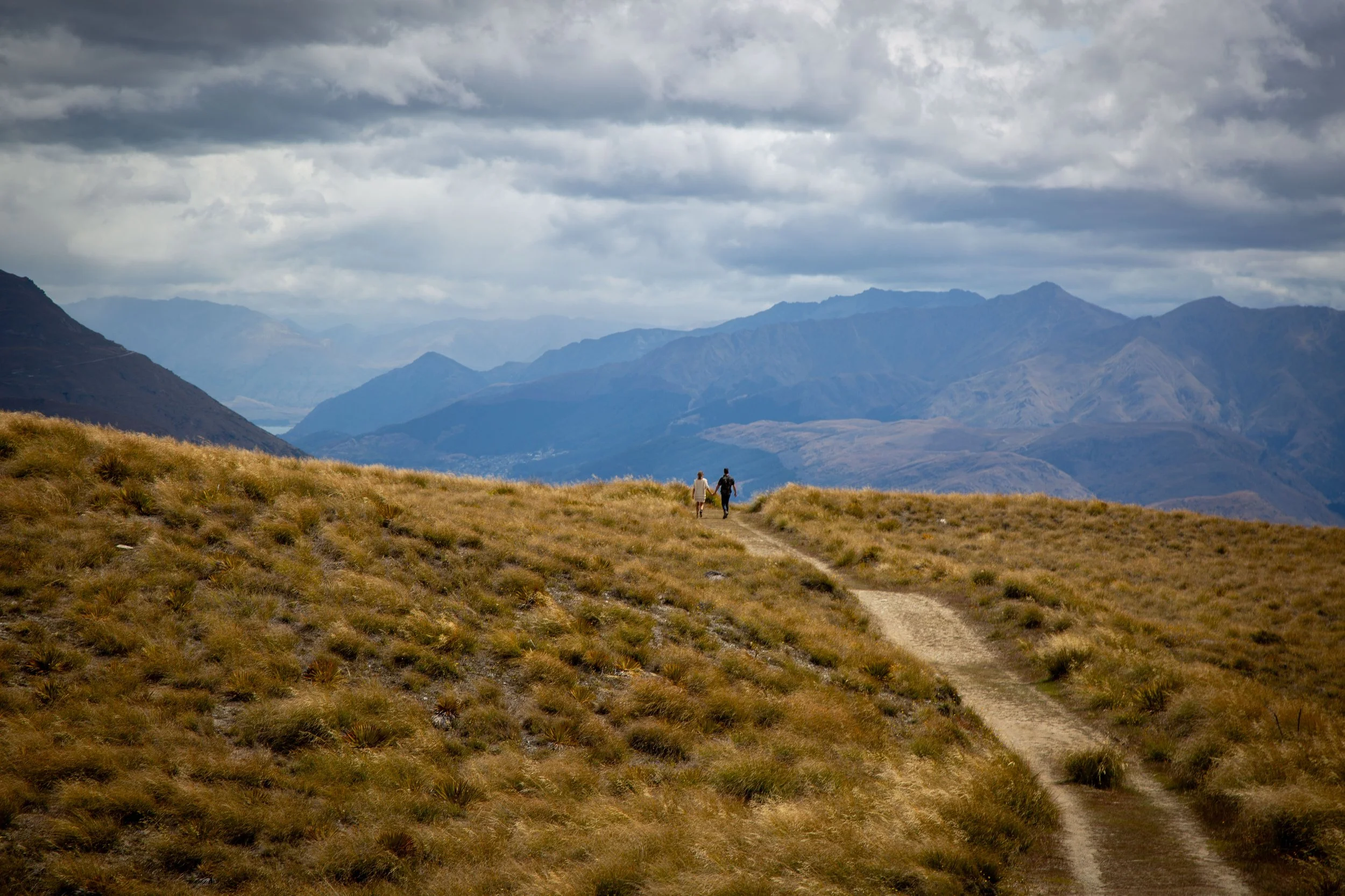 Two people walking on a dirt path through a grassy hillside with mountains in the background under cloudy skies.