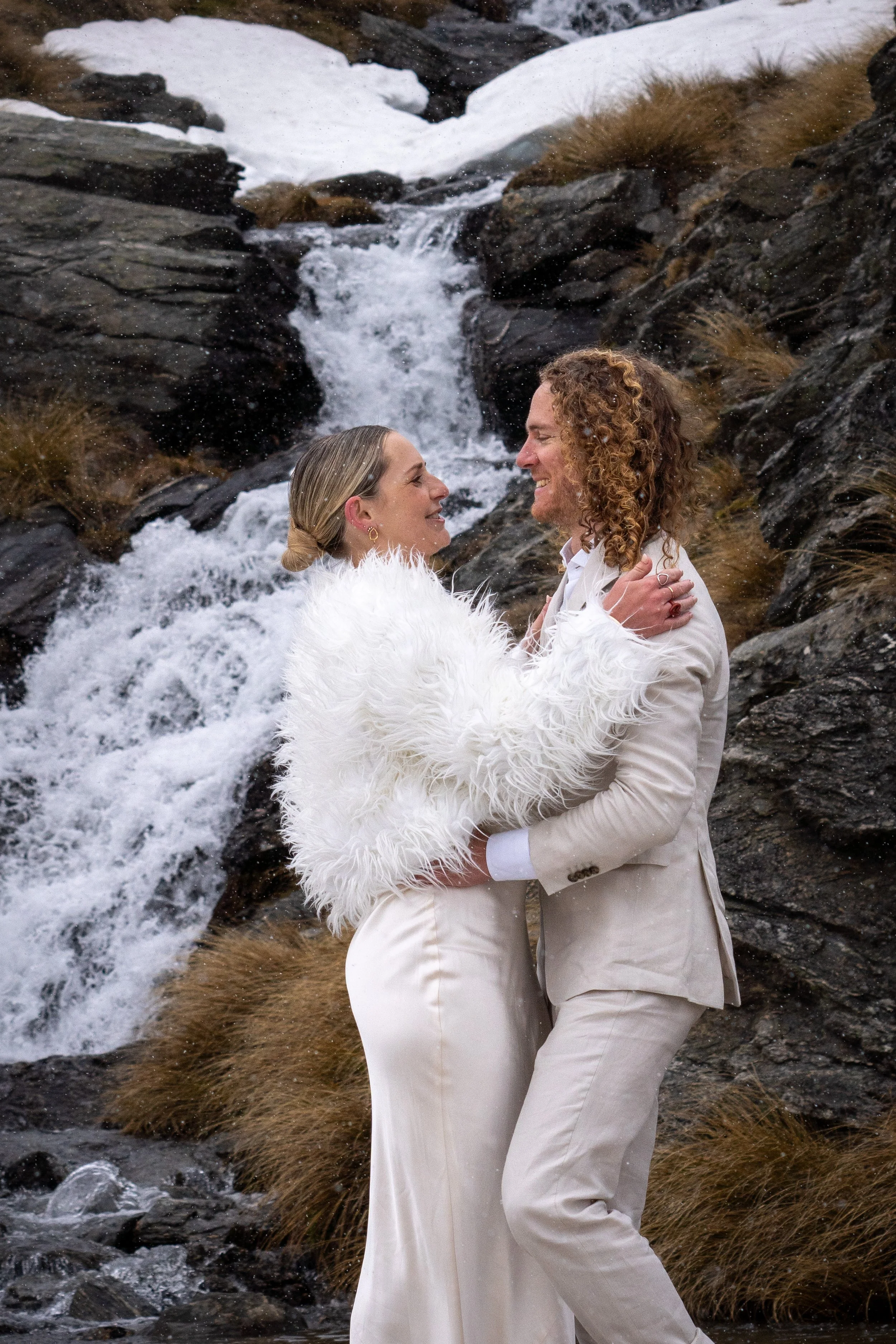 A couple in wedding attire embrace in front of a mountain creek with rocks, snow, and brown grass.