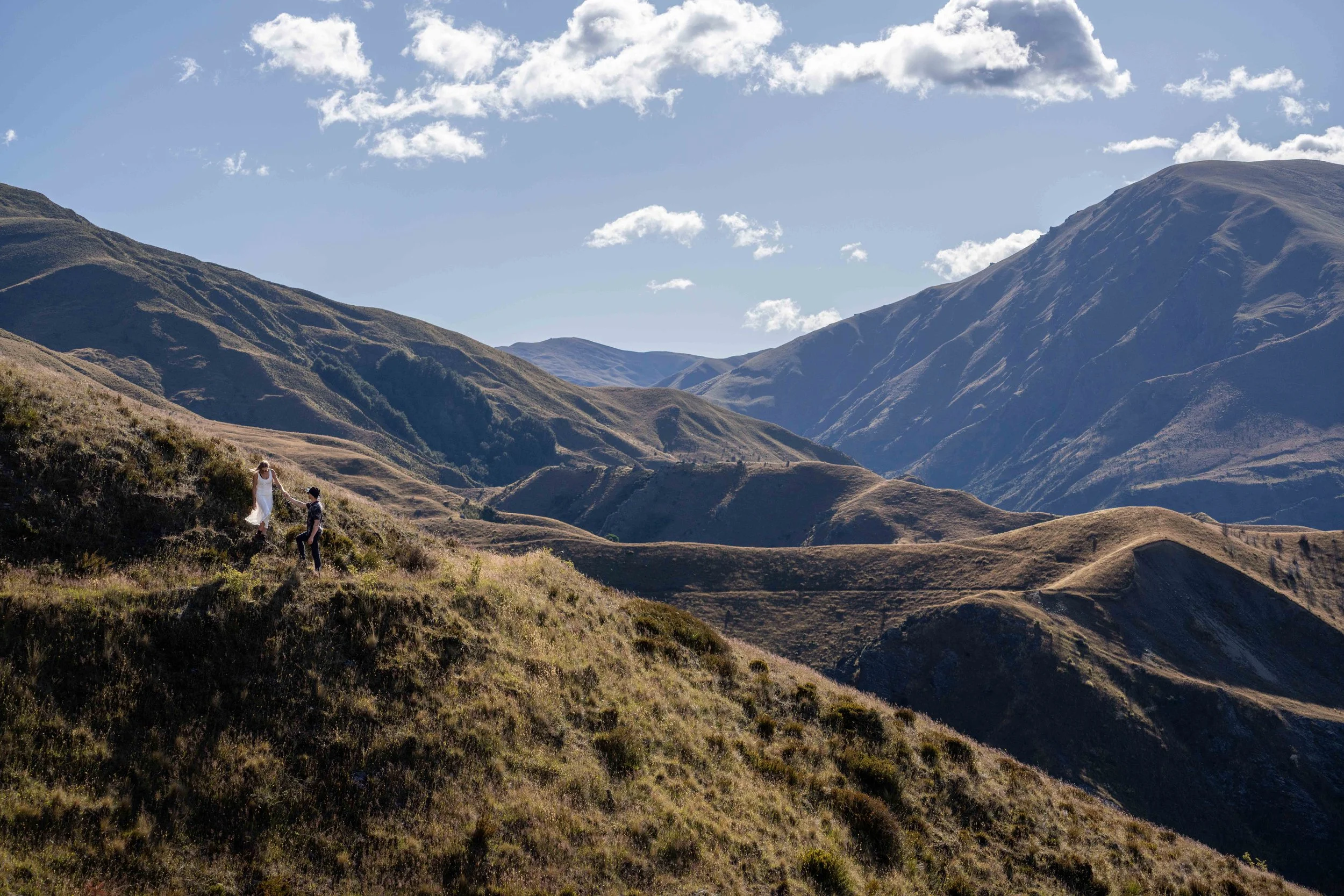 A couple walking in a grassy mountain landscape with rolling hills and peaks under a partly cloudy sky.