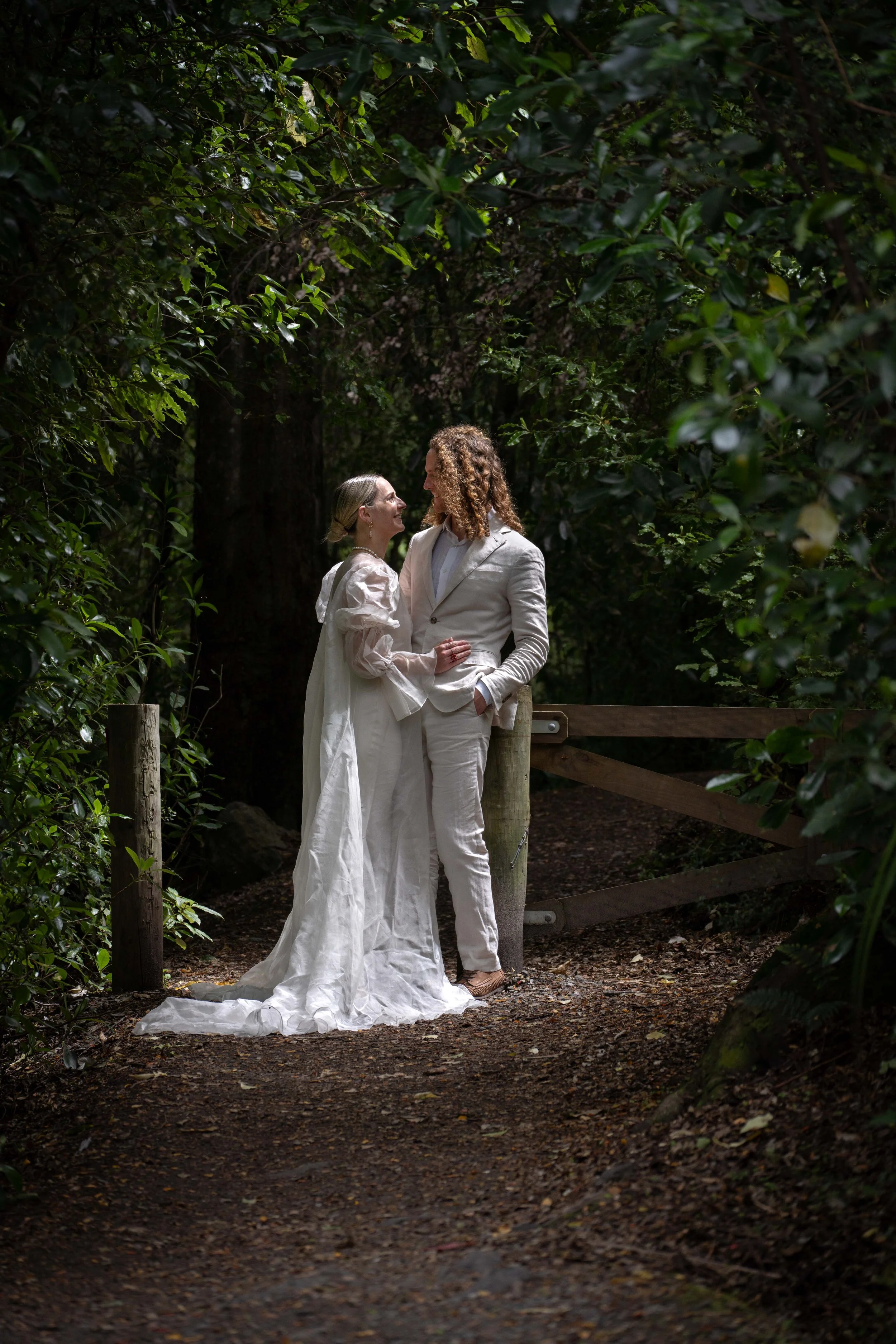 A couple in white wedding attire standing close on a forest path, gazing into each other's eyes among green foliage.