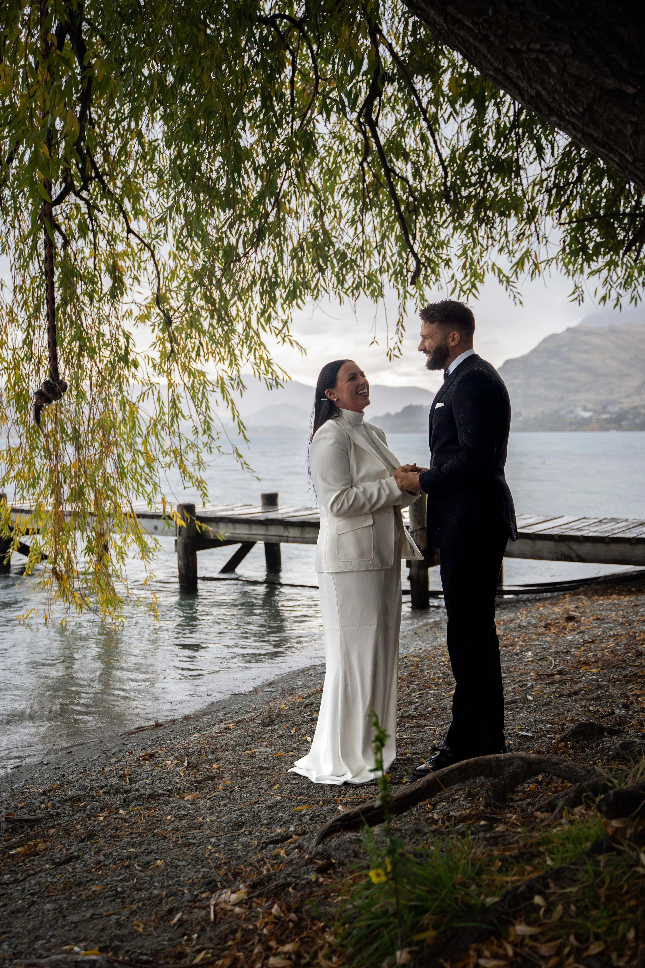 A bride and groom holding hands and smiling at each other under a tree by a lake with a dock and mountains in the background.