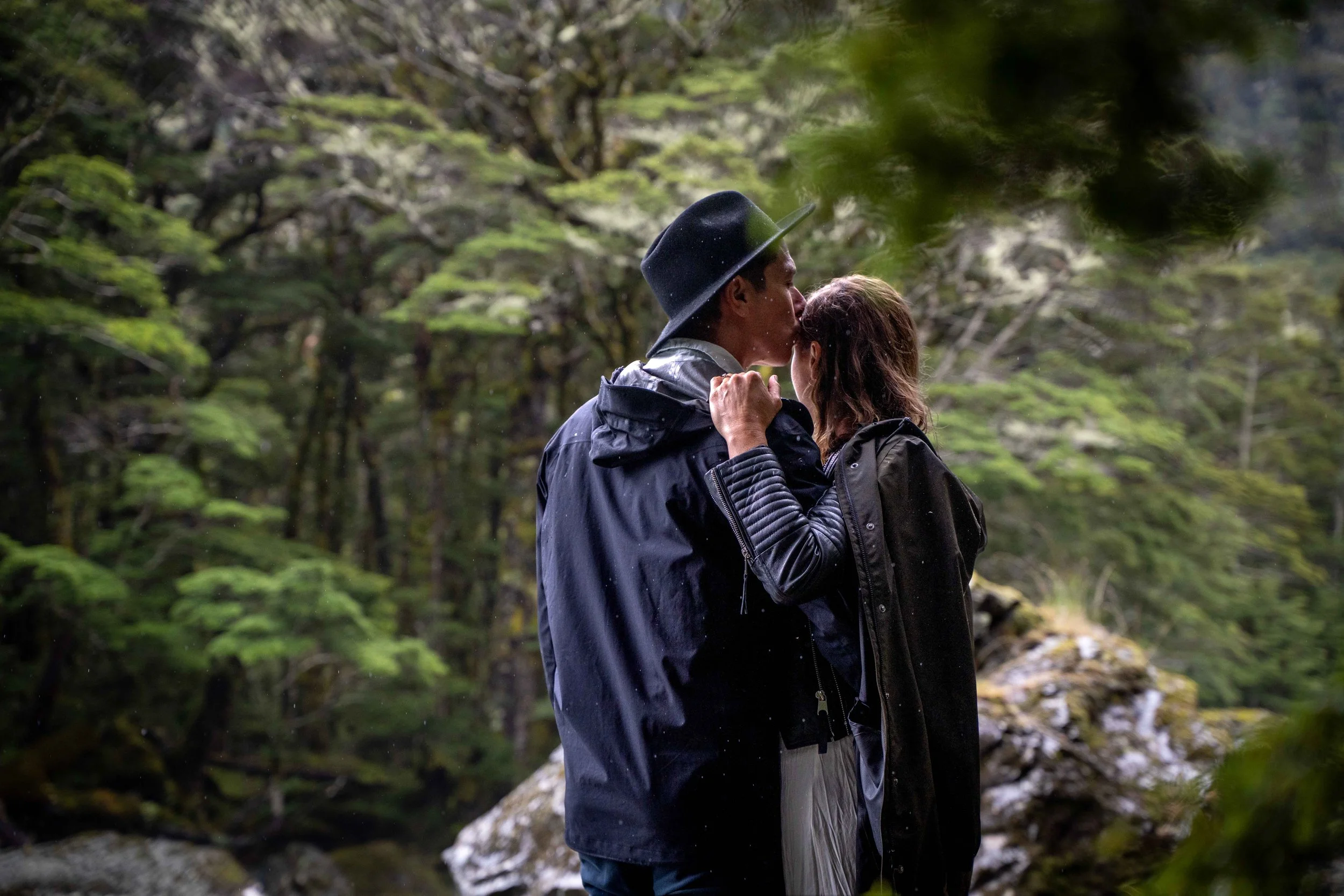 A couple embracing and kissing outdoors in a forested area, wearing rain jackets and a hat, with lush green trees and rocks in the background.