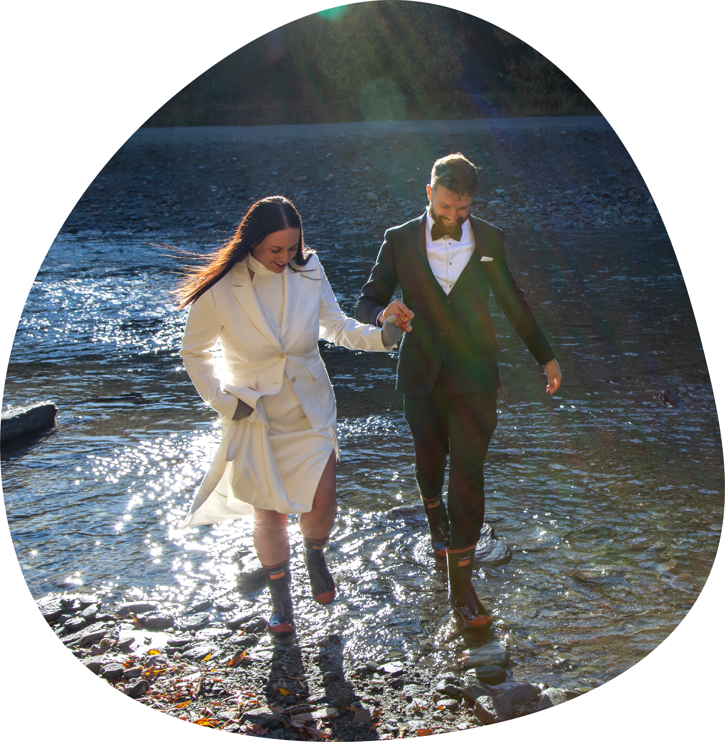 A couple dressed in formal wedding attire, holding hands and walking through a shallow river on a sunny day, with sunlight reflecting on the water.