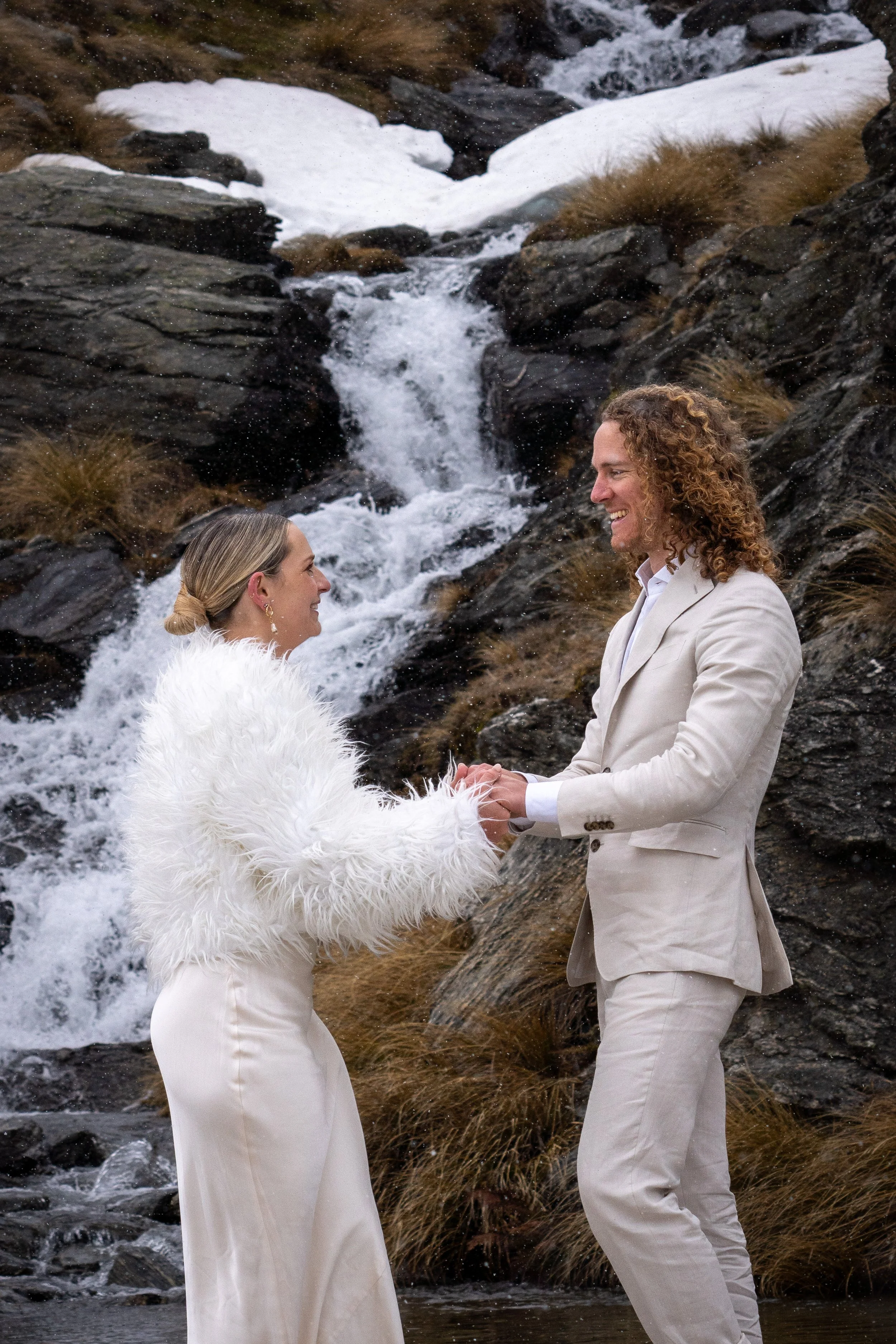 A couple dressed in formal wedding attire holding hands and smiling at each other in front of a mountain stream with small waterfalls and rocks.