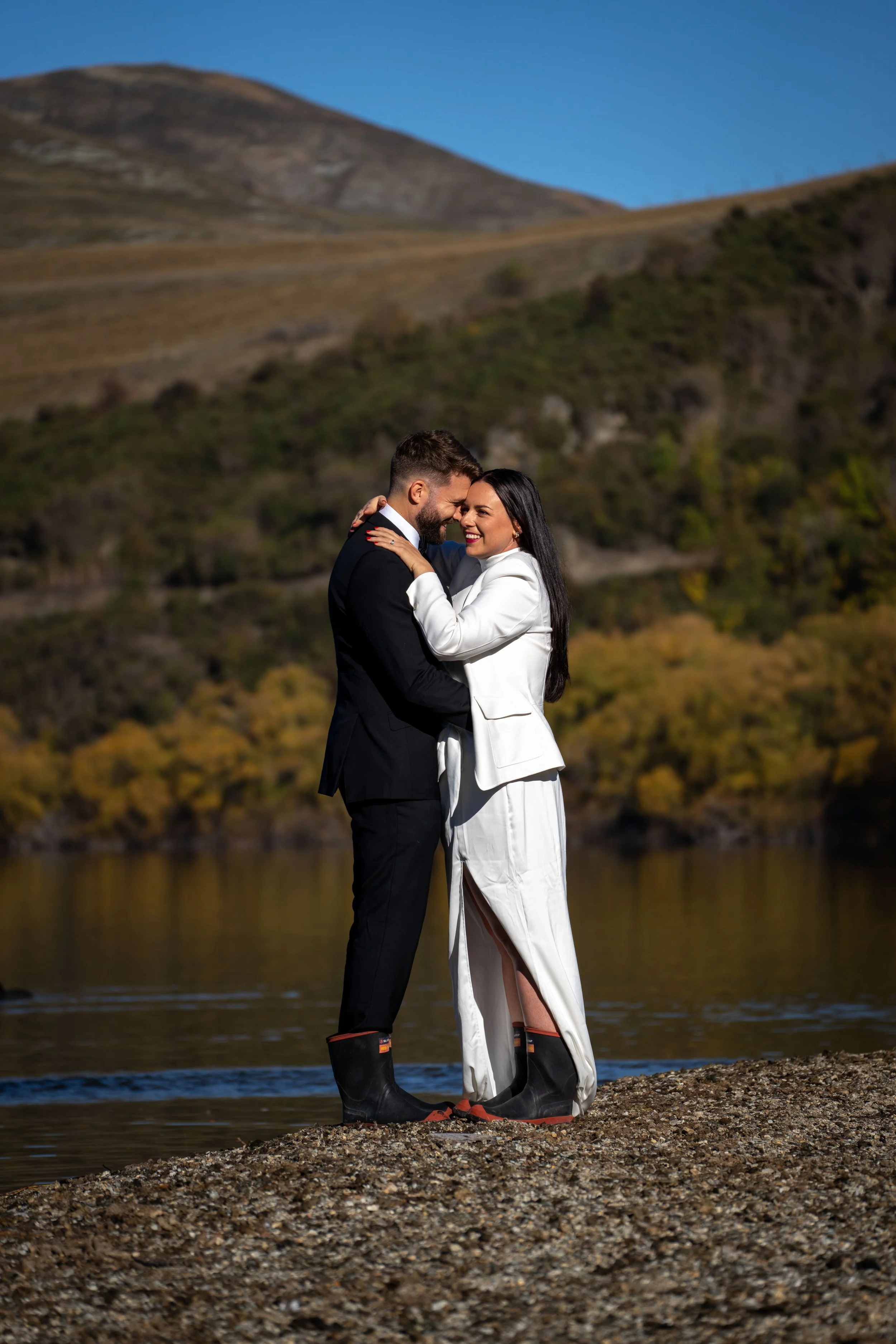 A couple in wedding attire smiling and embracing each other on a rocky riverbank with a scenic background of hills and trees.