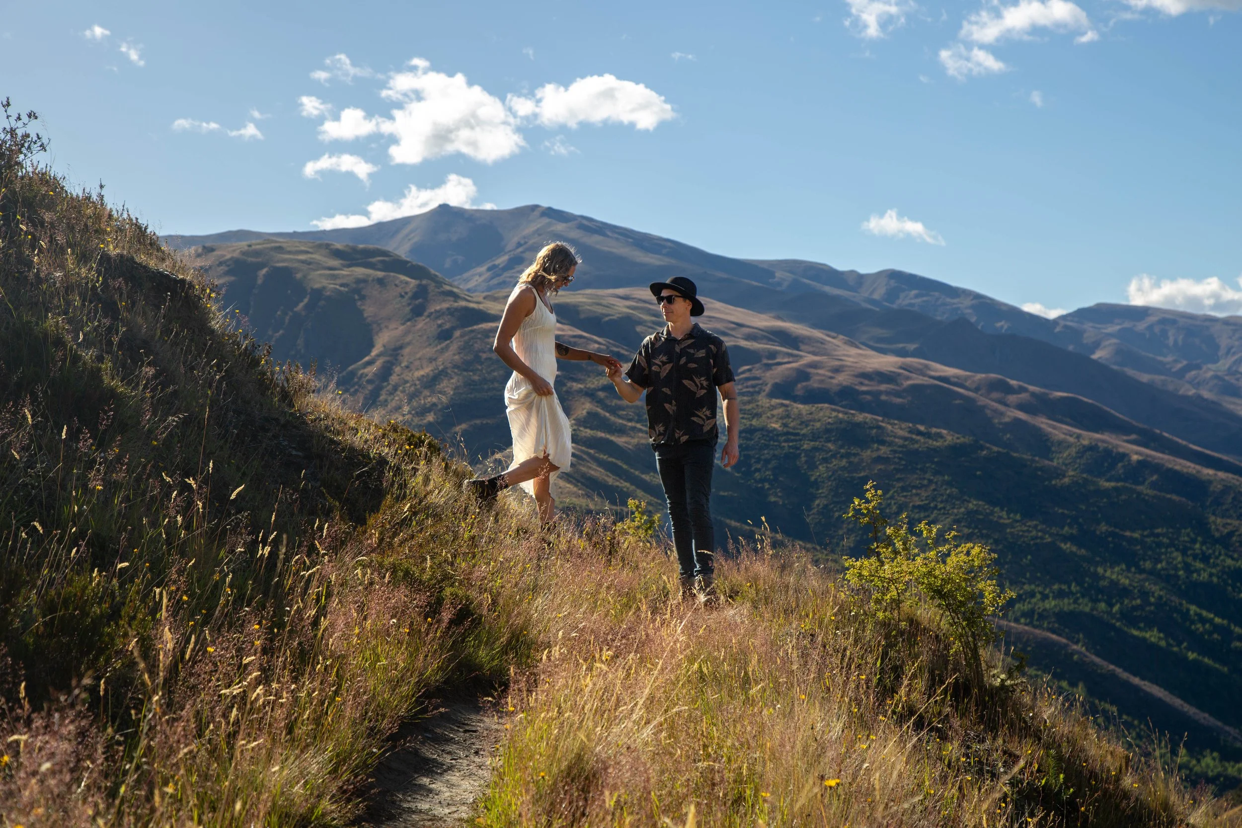 A man and woman on a mountain trail, with the man handing the woman a small object or flower, surrounded by rolling mountains and a blue sky with clouds.
