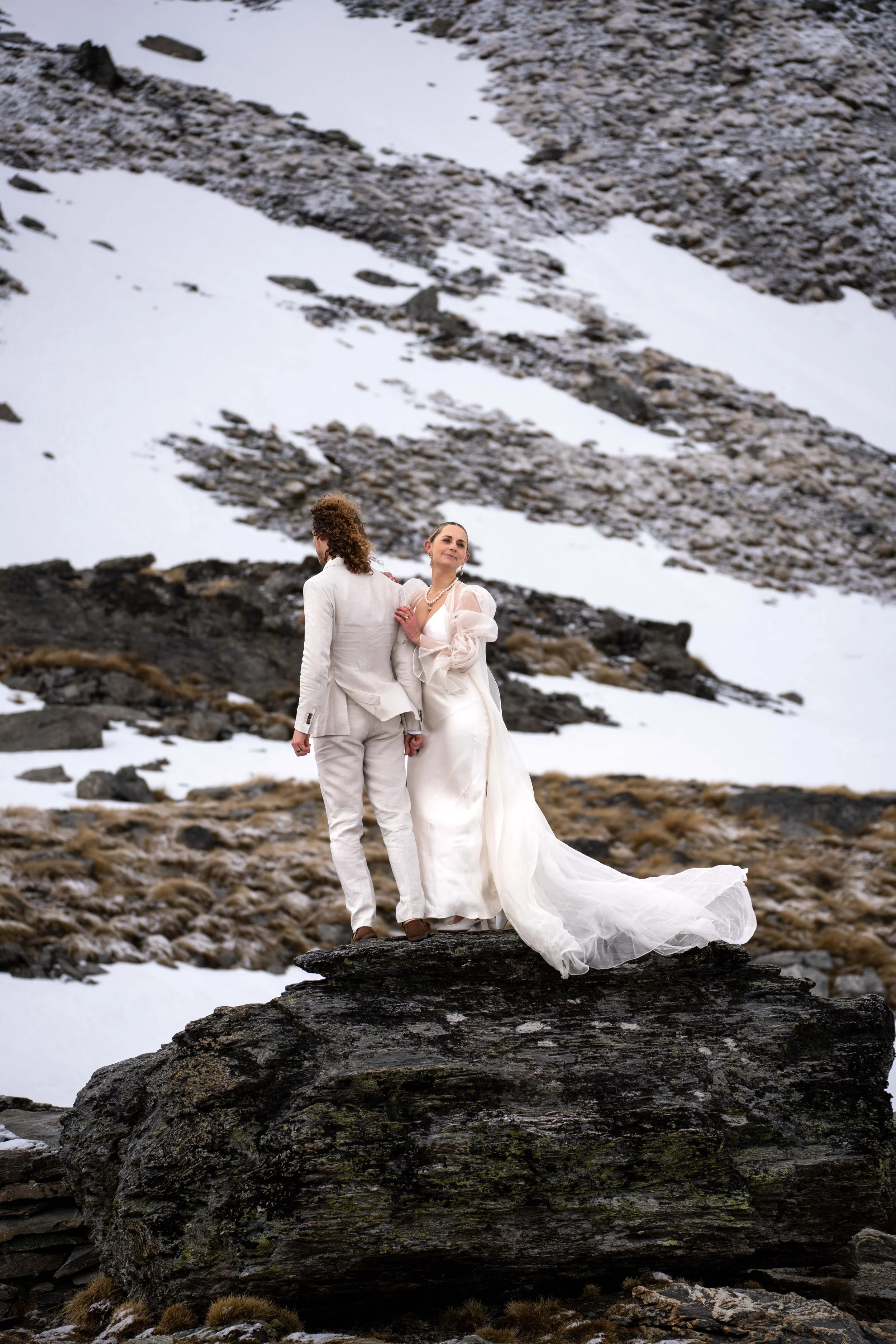 A bride and groom standing close together on a large rock in a mountainous area with snow and rocks in the background.