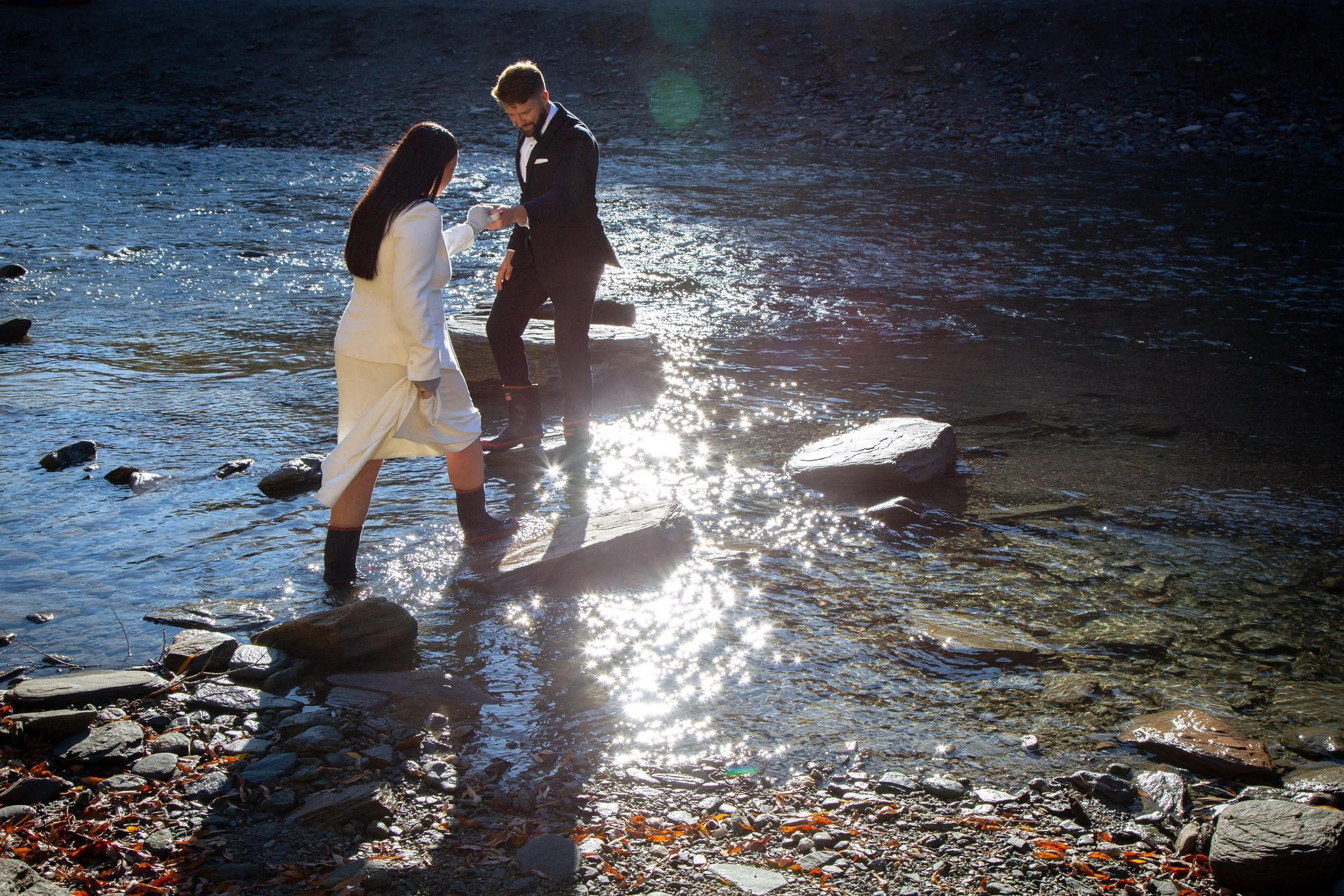 A couple getting married, standing in a river with rocks, with the bride in a white wedding dress and the groom in a black suit, holding hands and looking at each other.