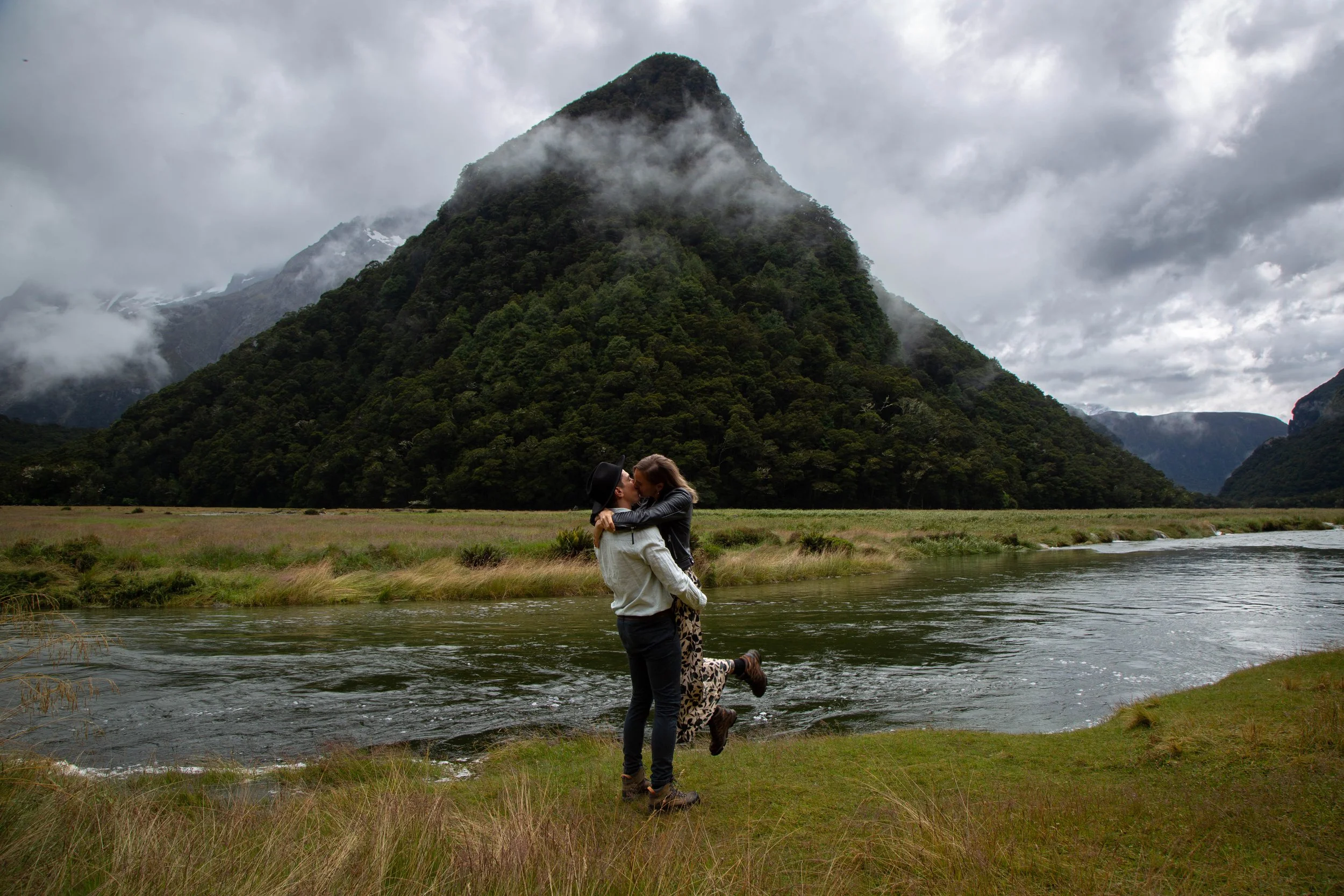 A couple sharing a kiss near a river with a lush green, fog-covered mountain in the background.