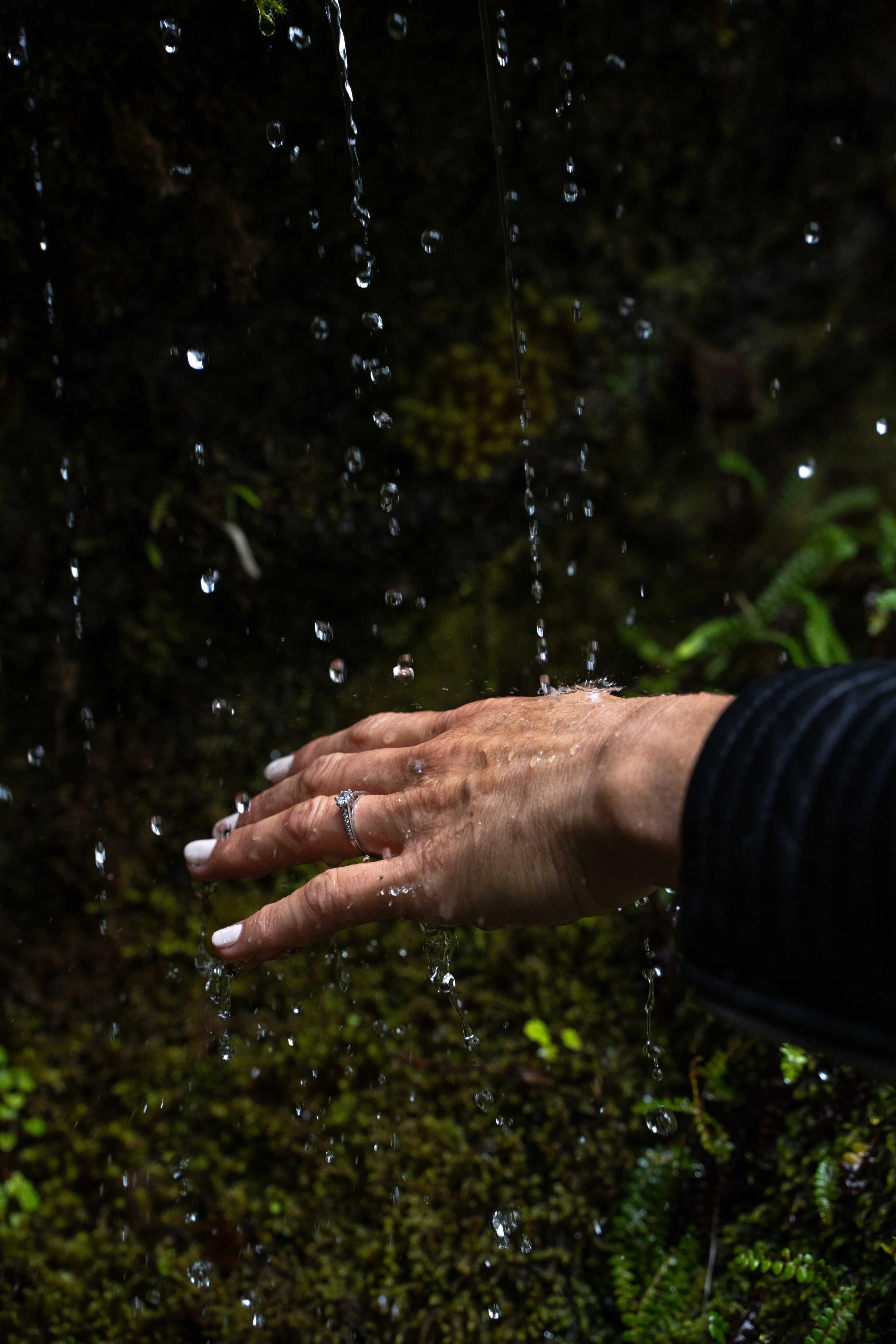 A hand reaching into a stream of falling water, with a ring on the finger and mossy ground in the background.