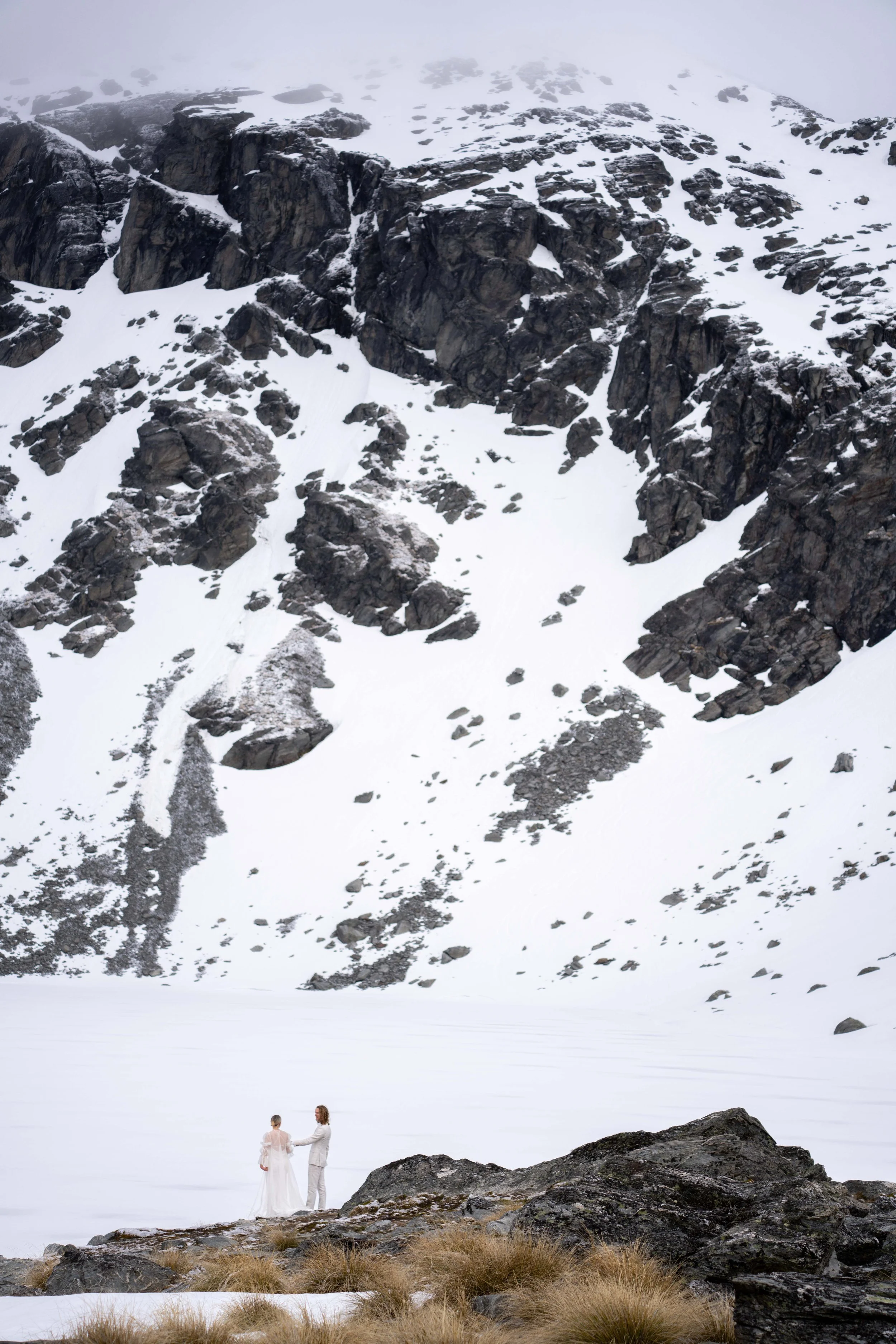 A couple dressed in wedding attire stand on rocky ground in front of a snowy mountain landscape.