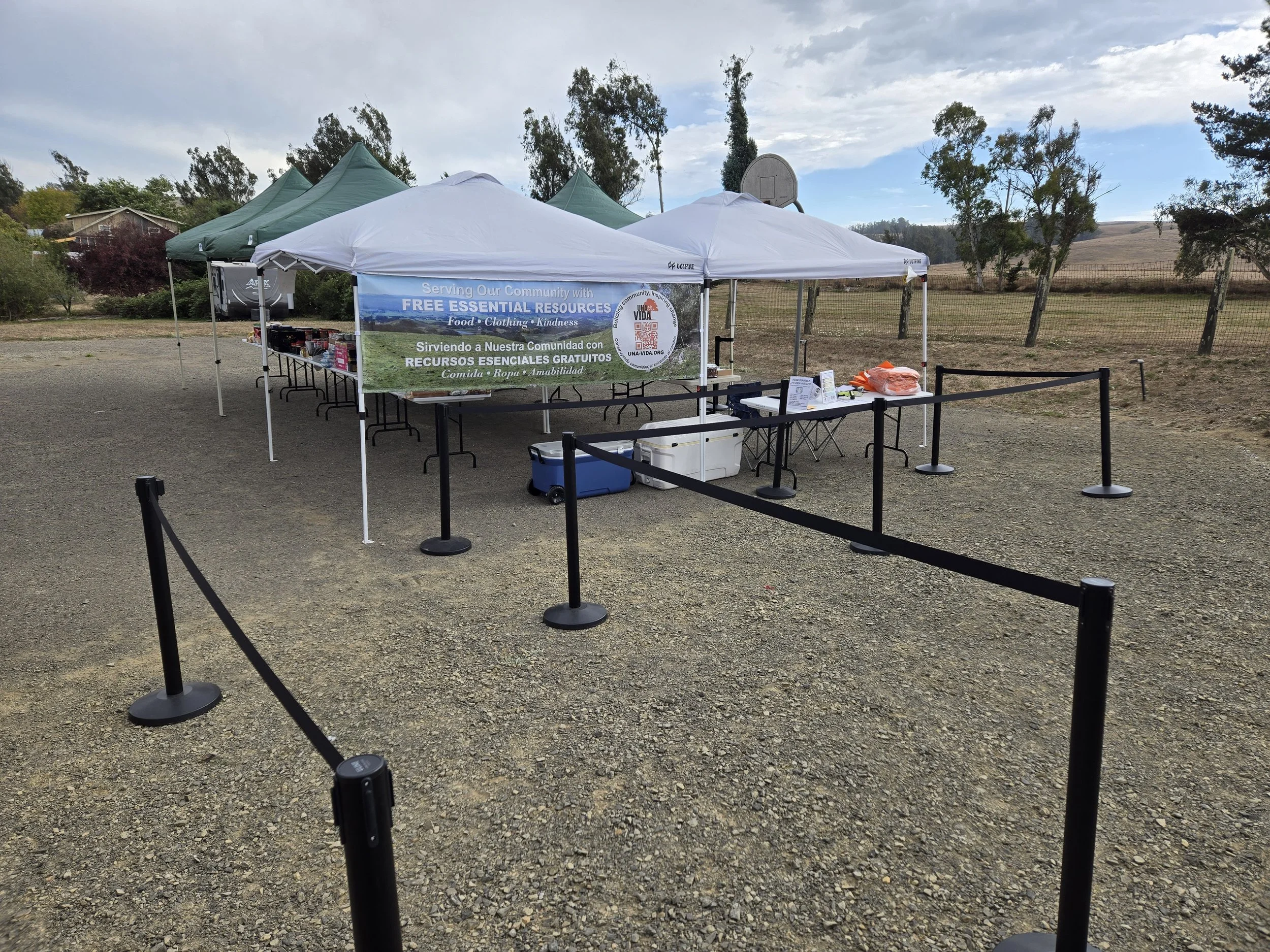 Una Vida’s outdoor food distribution in rural Valley Ford, with canopy tents, tables of free groceries, and a bilingual Una Vida banner surrounded by the Sonoma County countryside.