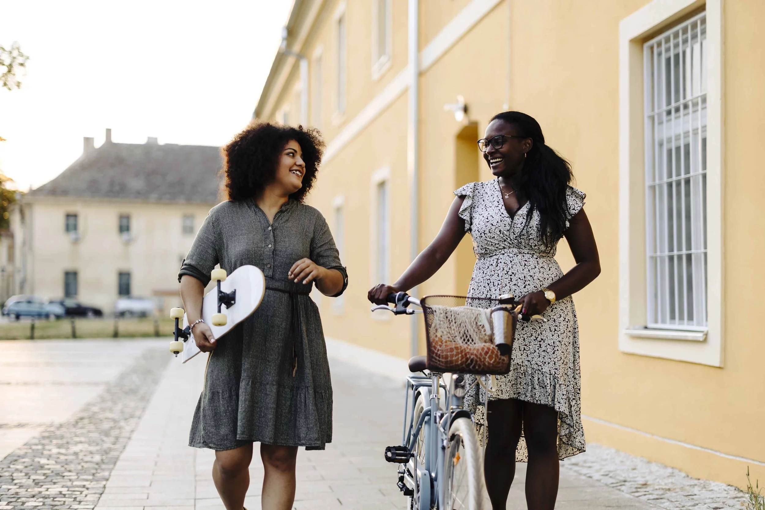 Two women walking and talking outside on a sunny day, one holding a skateboard and the other with a bicycle, smiling at each other near a yellow building.