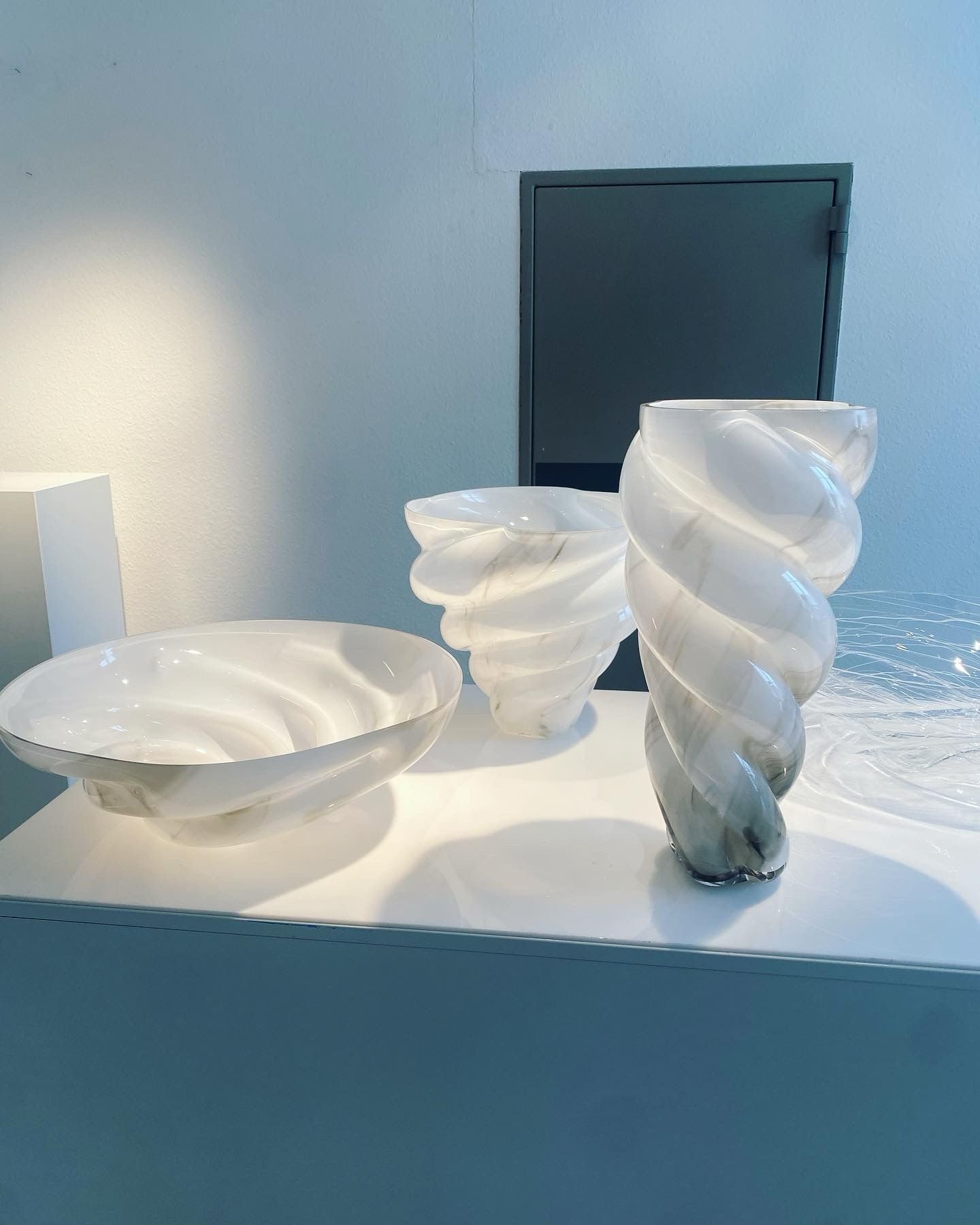 White abstract glass vases and bowls with swirling patterns on a white table against a light blue wall.