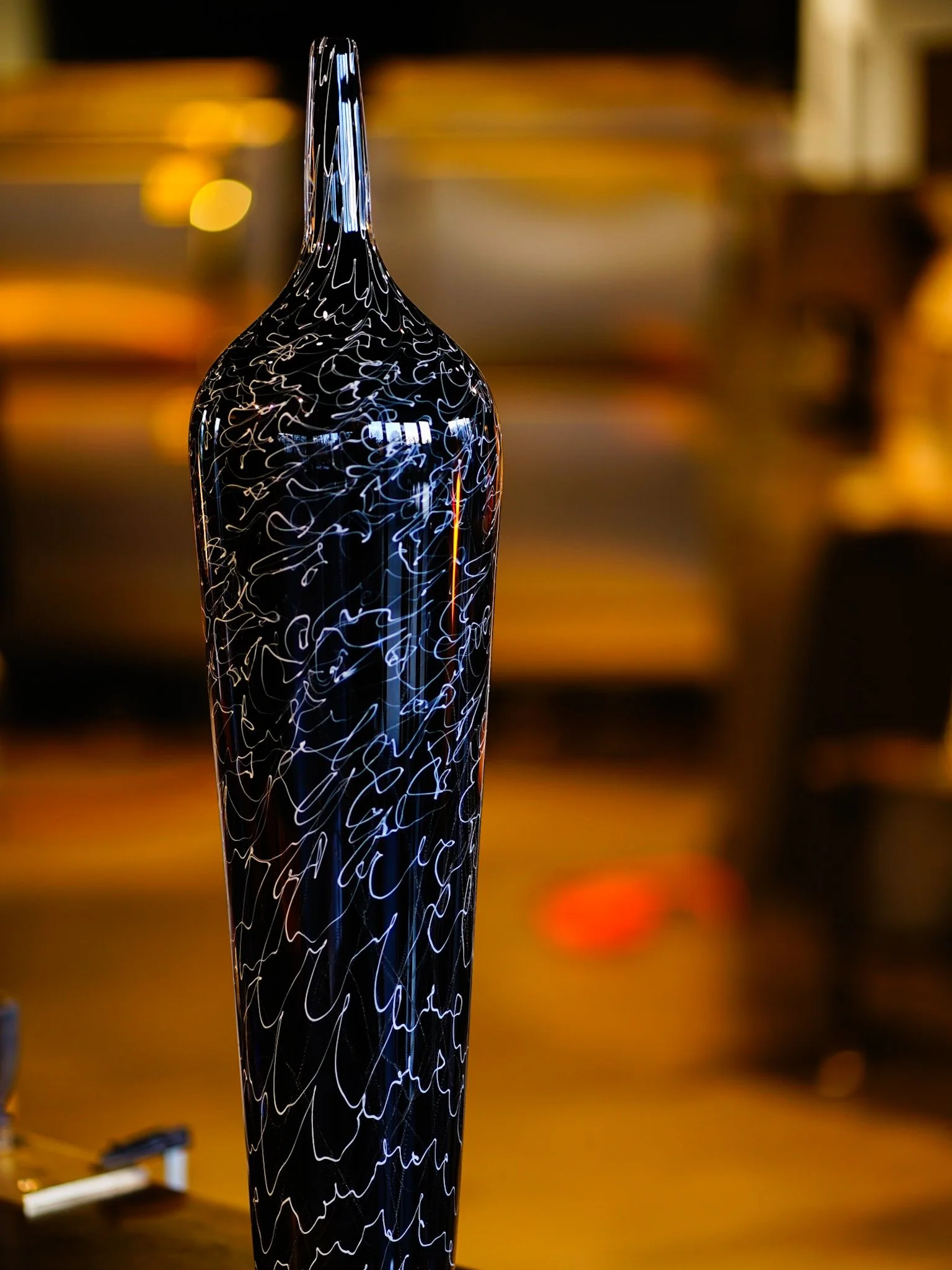 Close-up of a glass sculpture with black acrylic and white script design, set against a blurred warm-toned background.