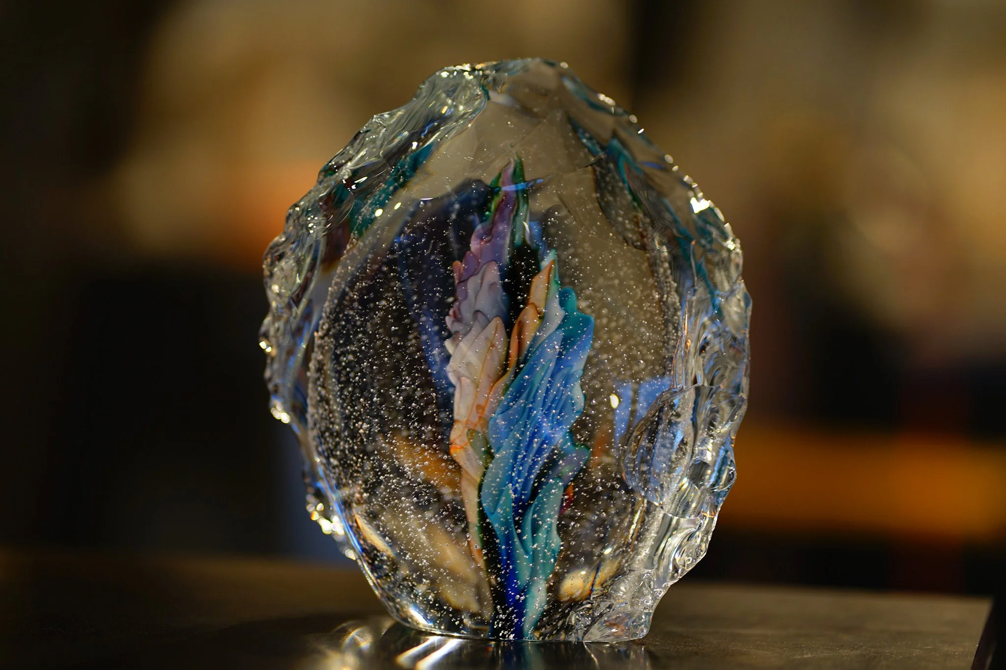A detailed close-up of an abstract glass sculpture with swirling colors inside, placed on a reflective surface with a blurred background.