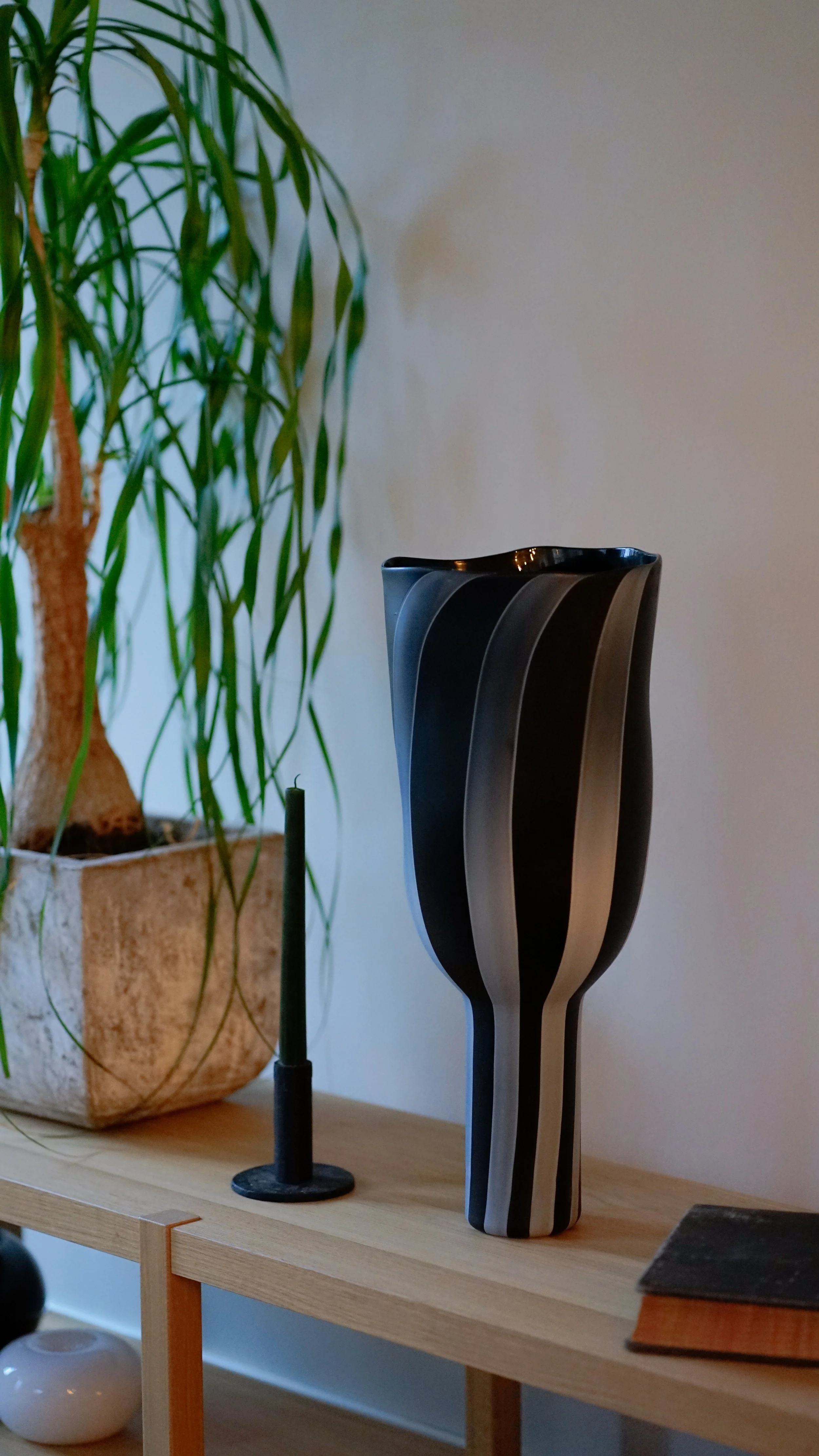 A wooden table with a tall black and gray striped ceramic vase, a black candlestick holder with a green candle, a white rounded vase, a potted plant with long green leaves, and a closed book.