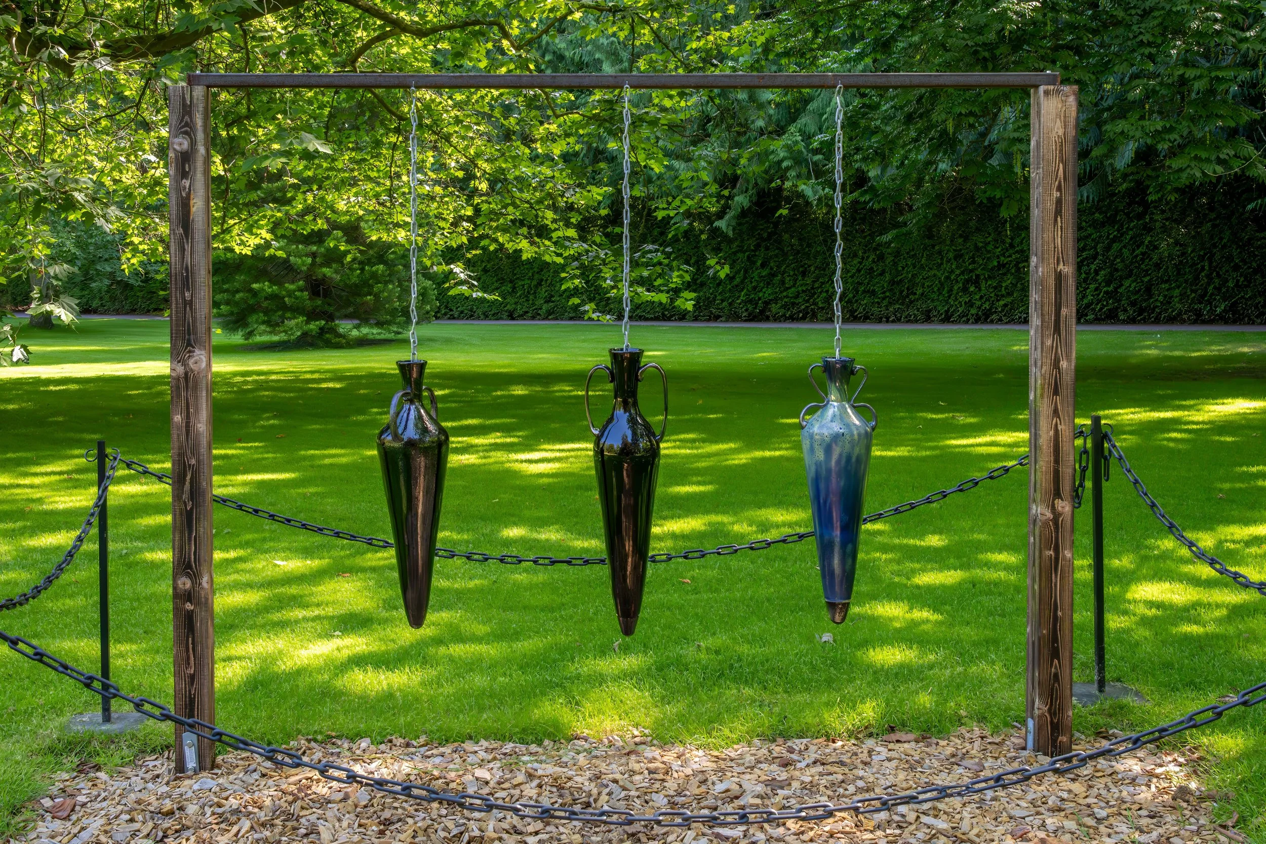 Four elongated glass vases hanging from a wooden frame with chain links, set outdoors on a grassy area with trees in the background.