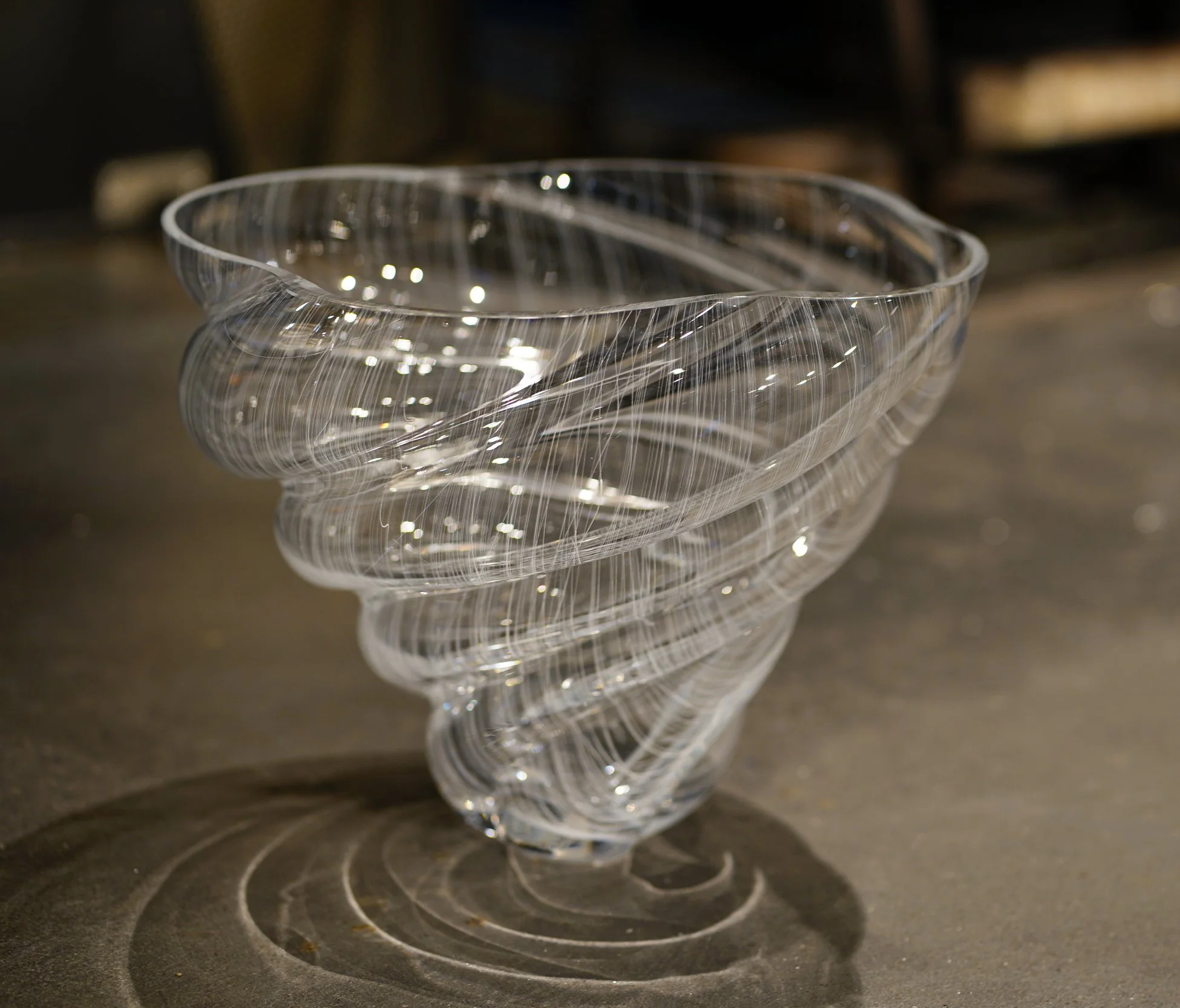 Close-up of an elegant, clear glass bowl with thin, swirling designs, sitting on a dark surface with concentric circular patterns.