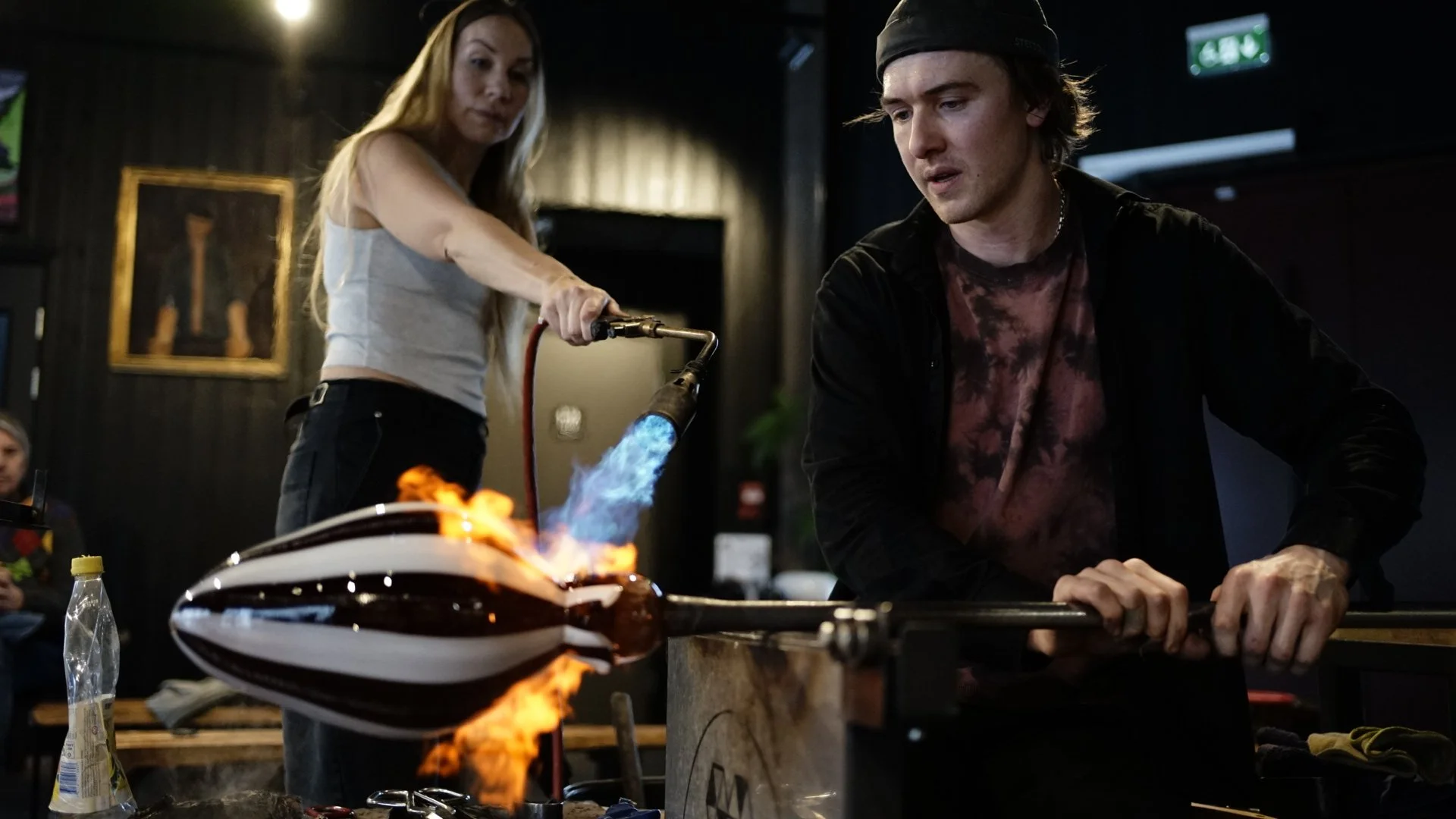 A woman with long blonde hair holding a blowtorch with a blue flame, appearing to melt or shape glass, while a young man with dark hair and a hat sits nearby at a table. In the background, there are dark walls, framed artwork, and a green exit sign.
