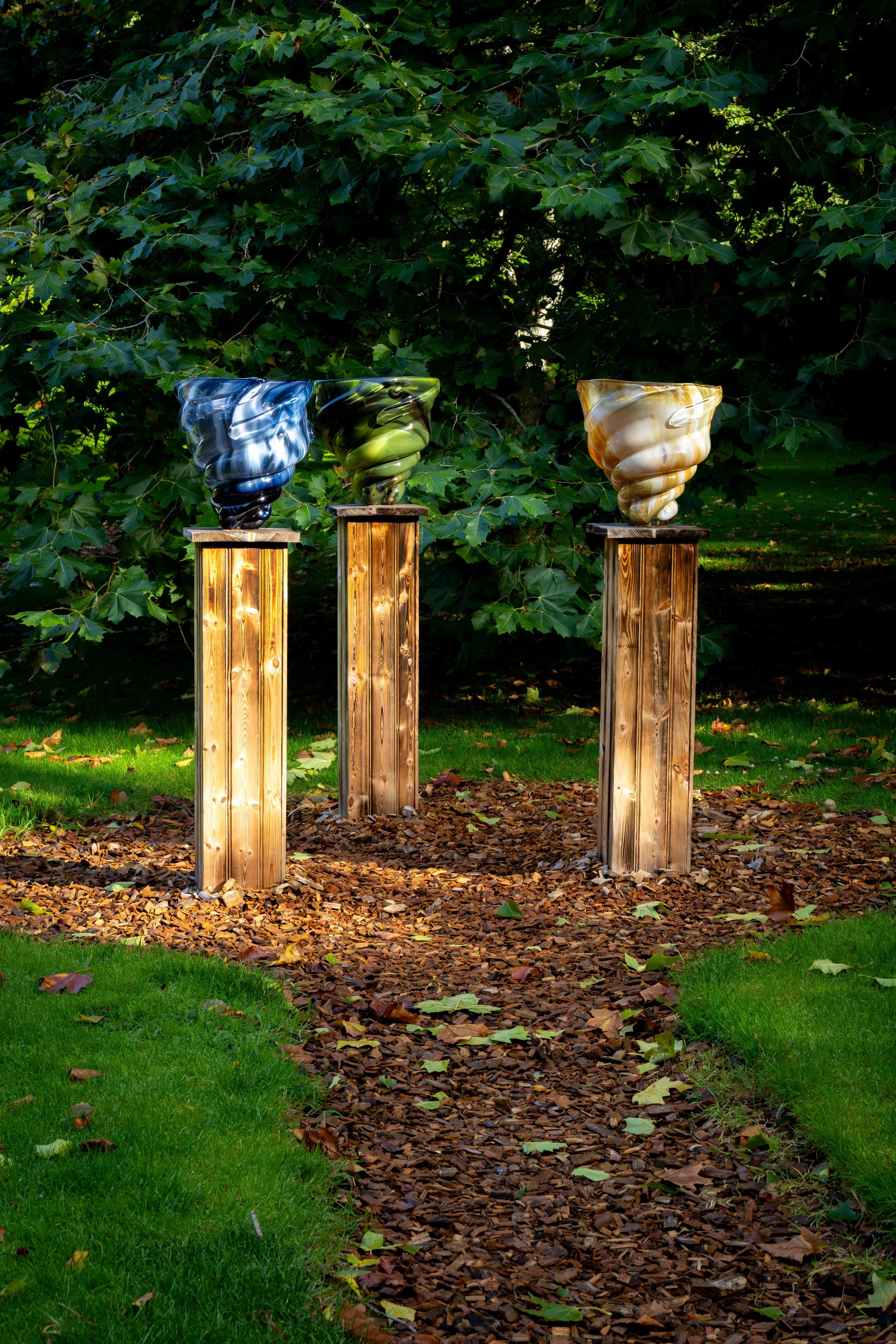 Three decorative glass vases with swirling patterns on wooden pedestals, set outdoors among green trees and fallen leaves.