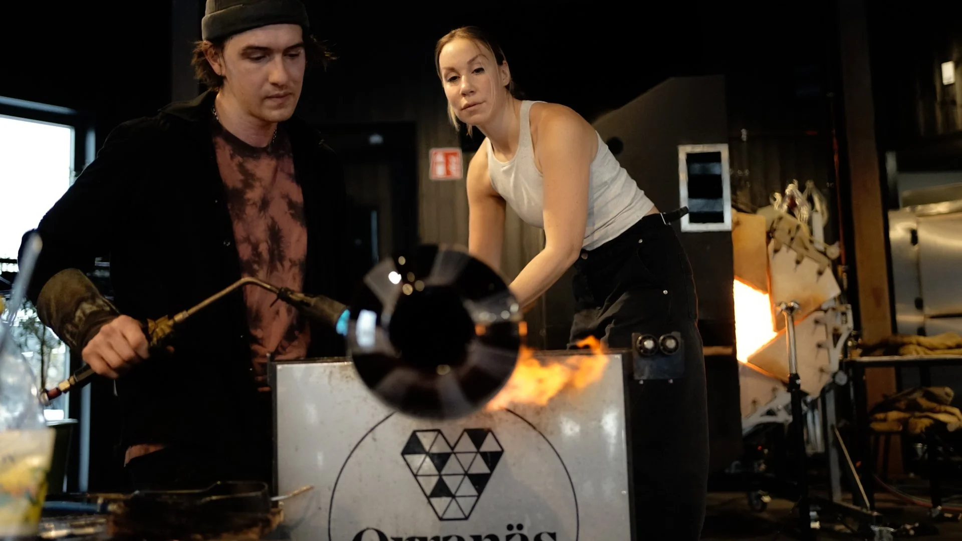 Two people operating a glassblowing furnace, one using a blowpipe and the other assisting, in a workshop.