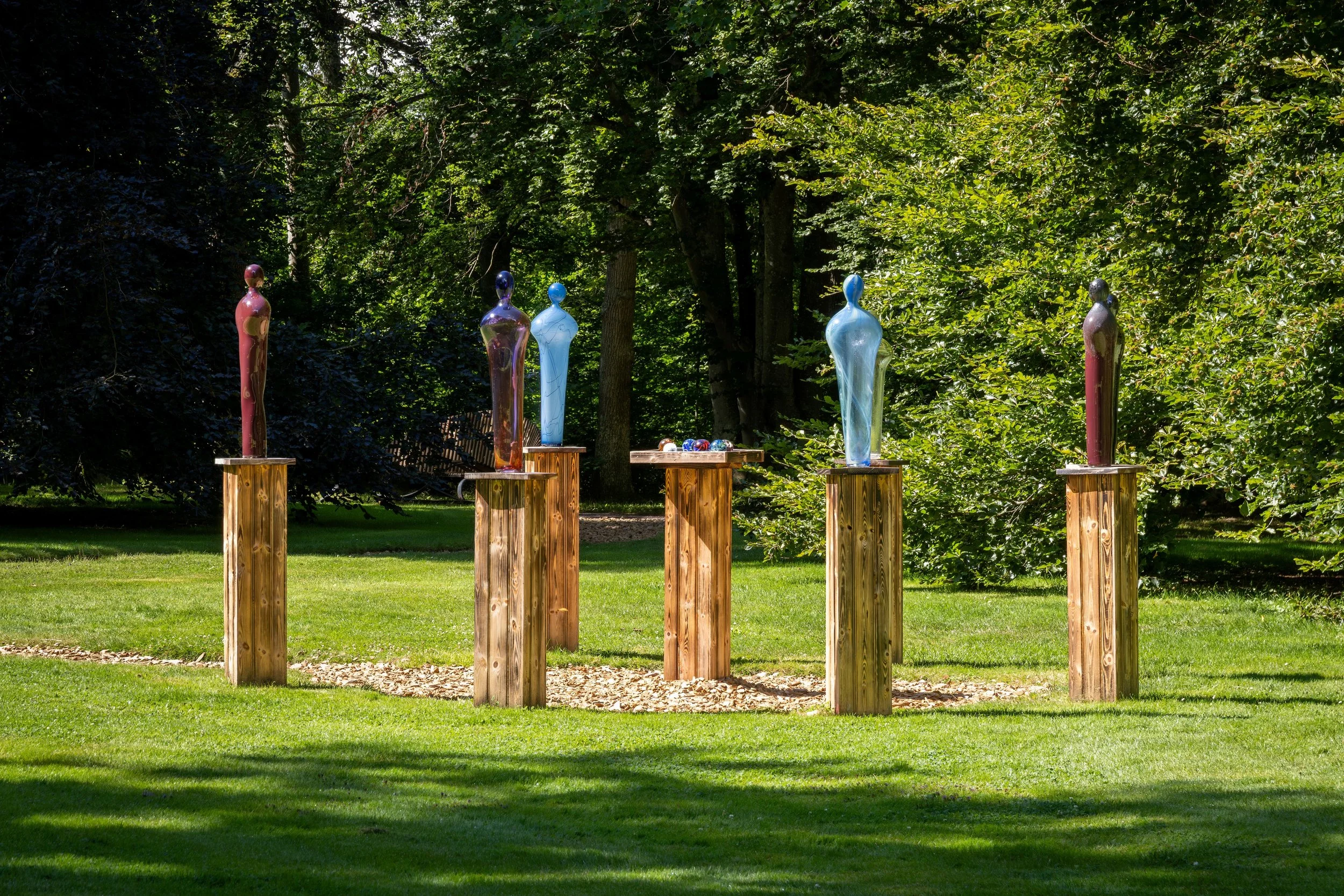Outdoor sculpture installation featuring several glass human figures in shades of pink, purple, blue, and gray, displayed on wooden pedestals in a garden setting with lush green trees in the background.