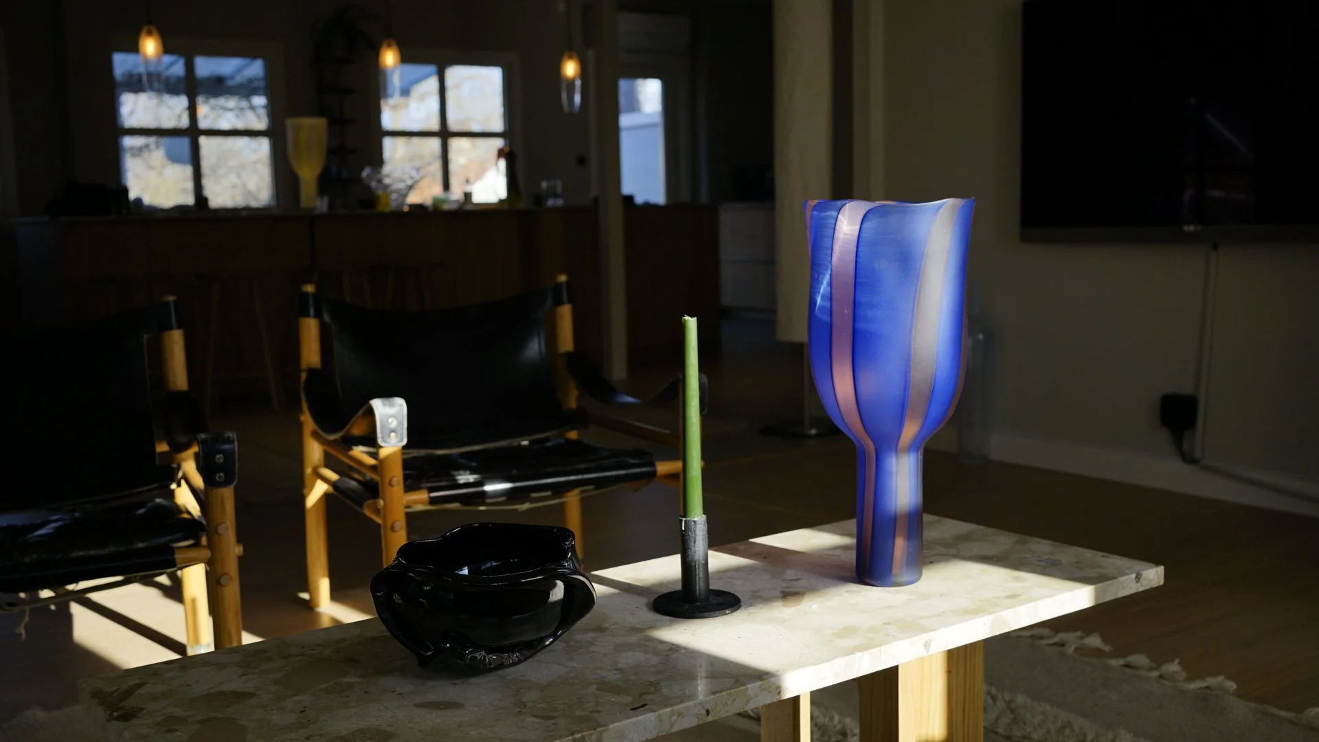 Interior of a house with sunlight streaming in, featuring a marble table with colorful glass art vases and bowls, black chairs, and a background of windows and pendant lights.