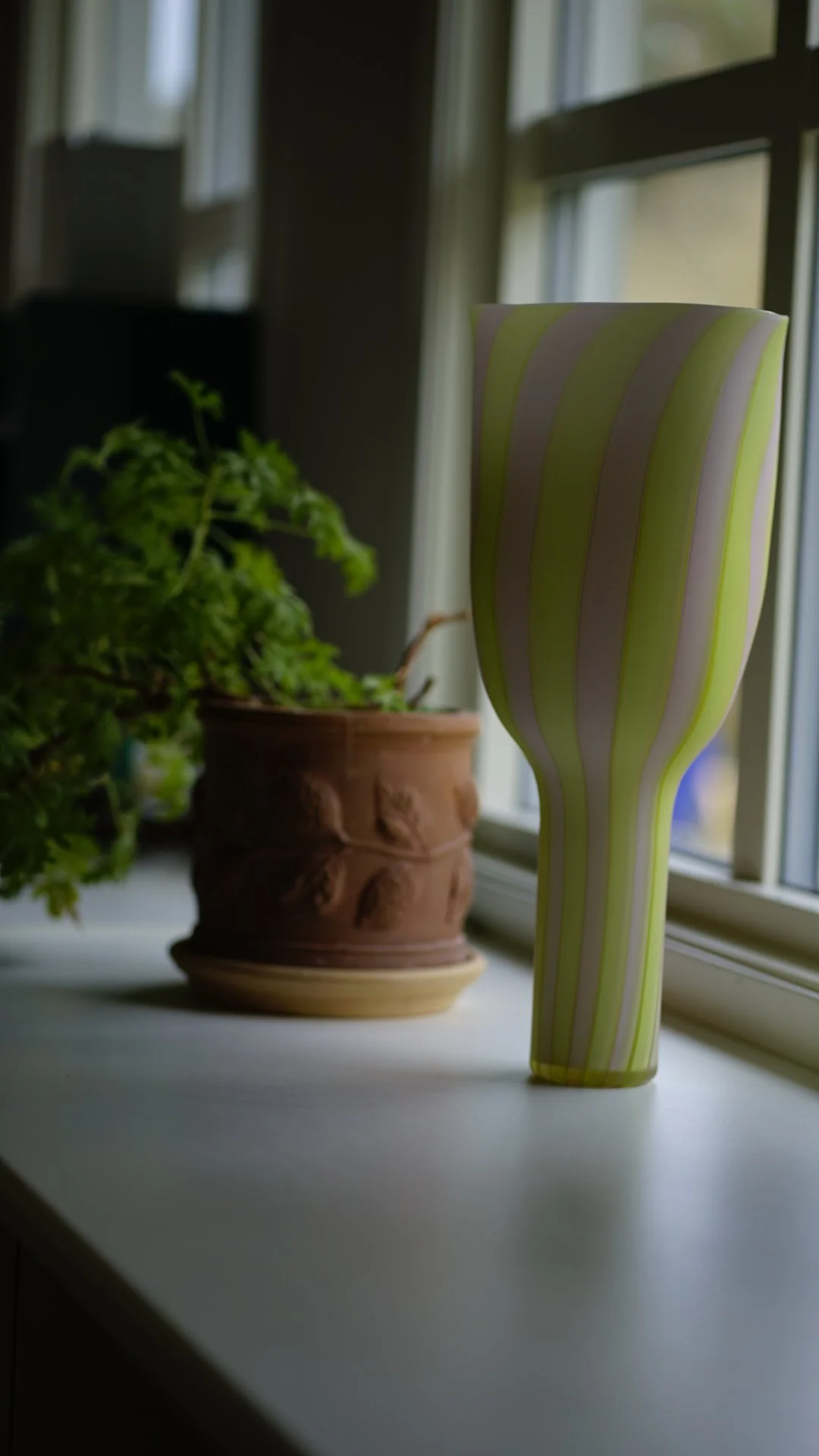 Close-up of a green and yellow striped vase on a windowsill next to a potted plant with green foliage.